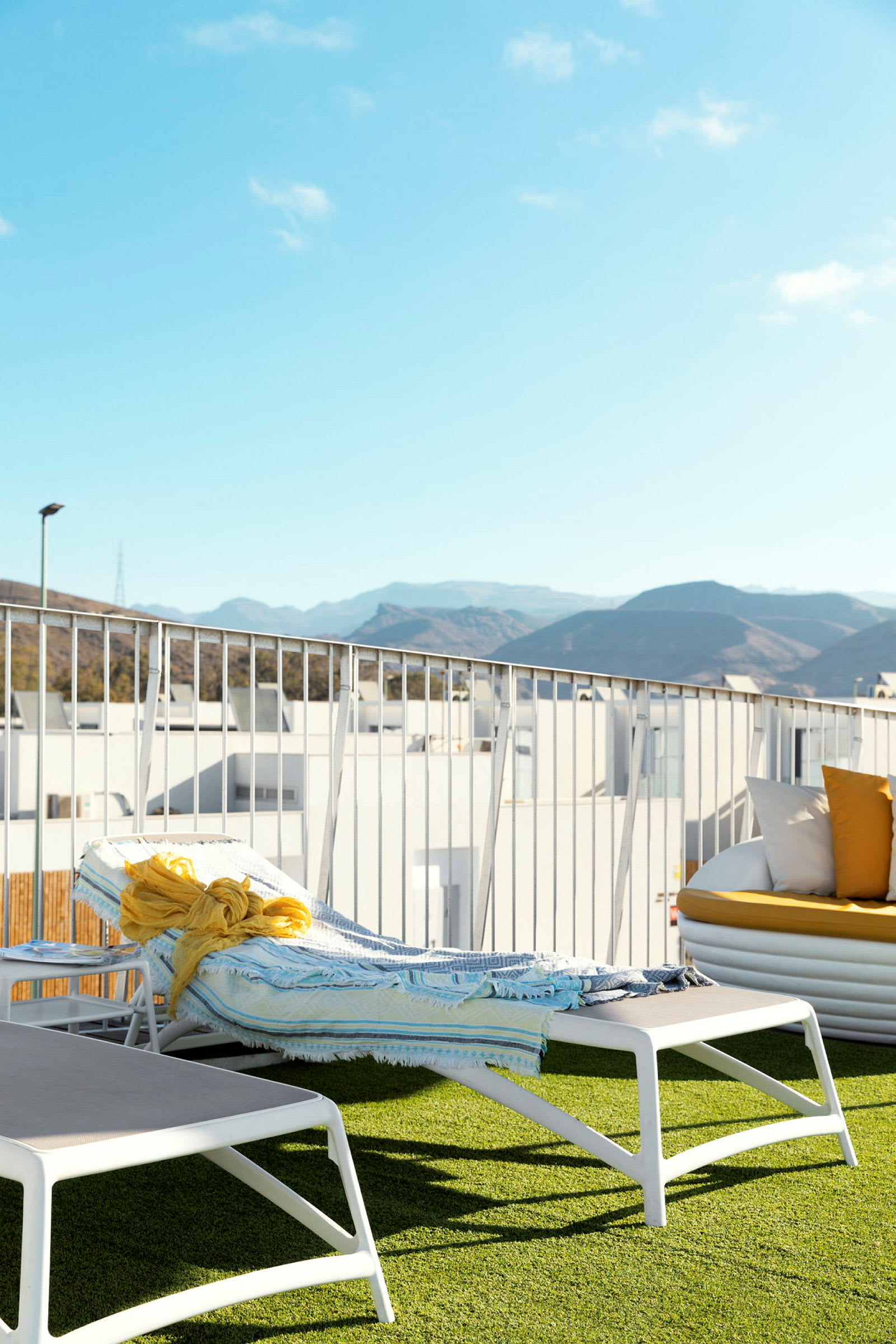 En solseng med et håndklæde og et gult sjal ligger på en terrasse med kunstgræs, med bjerge og blå himmel i baggrunden. (AI-genereret tekst)