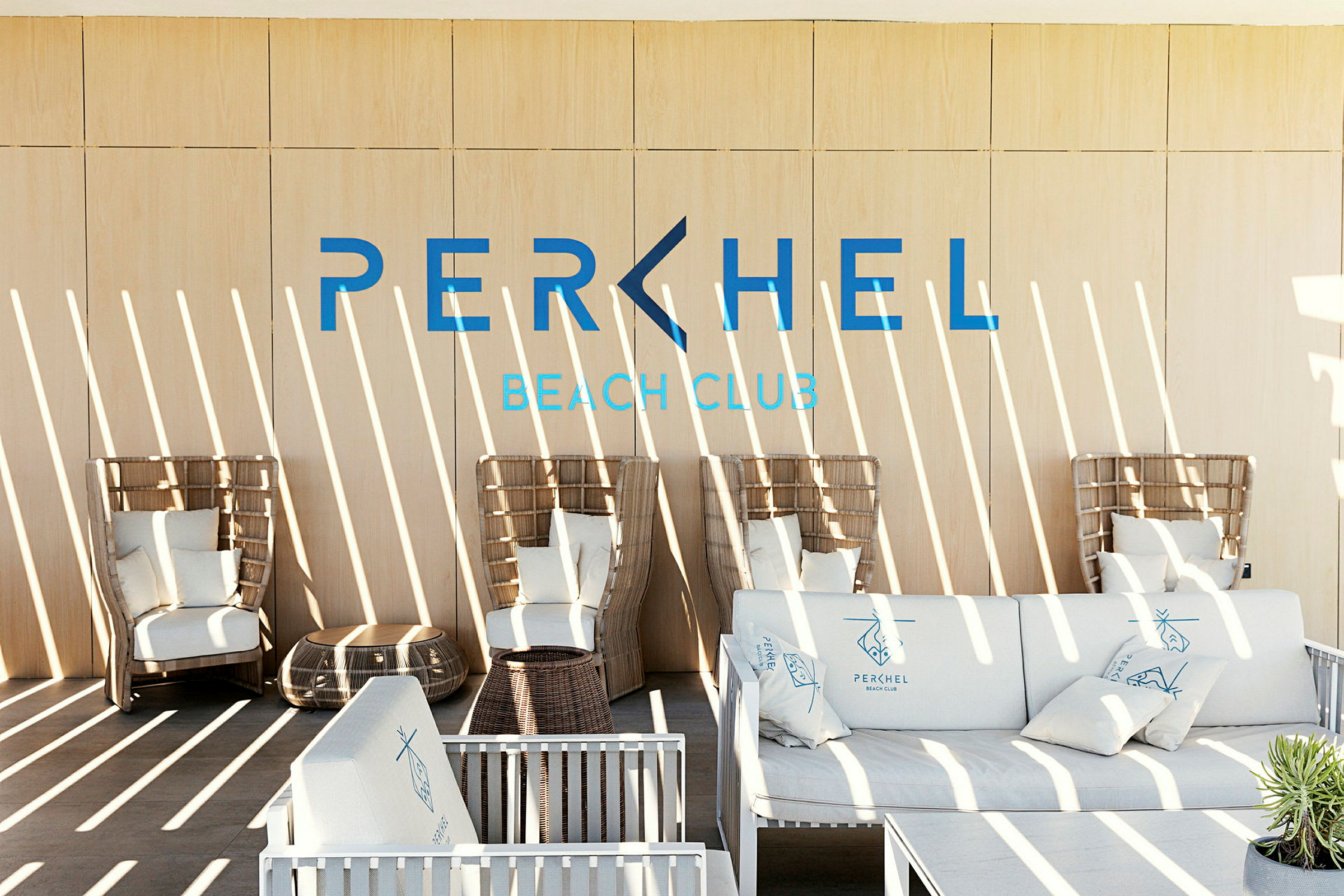 Et moderne loungeområde med kurvemøbler og sofaer med puder samt teksten 'Perchel Beach Club' på væggen. (AI-genereret tekst)