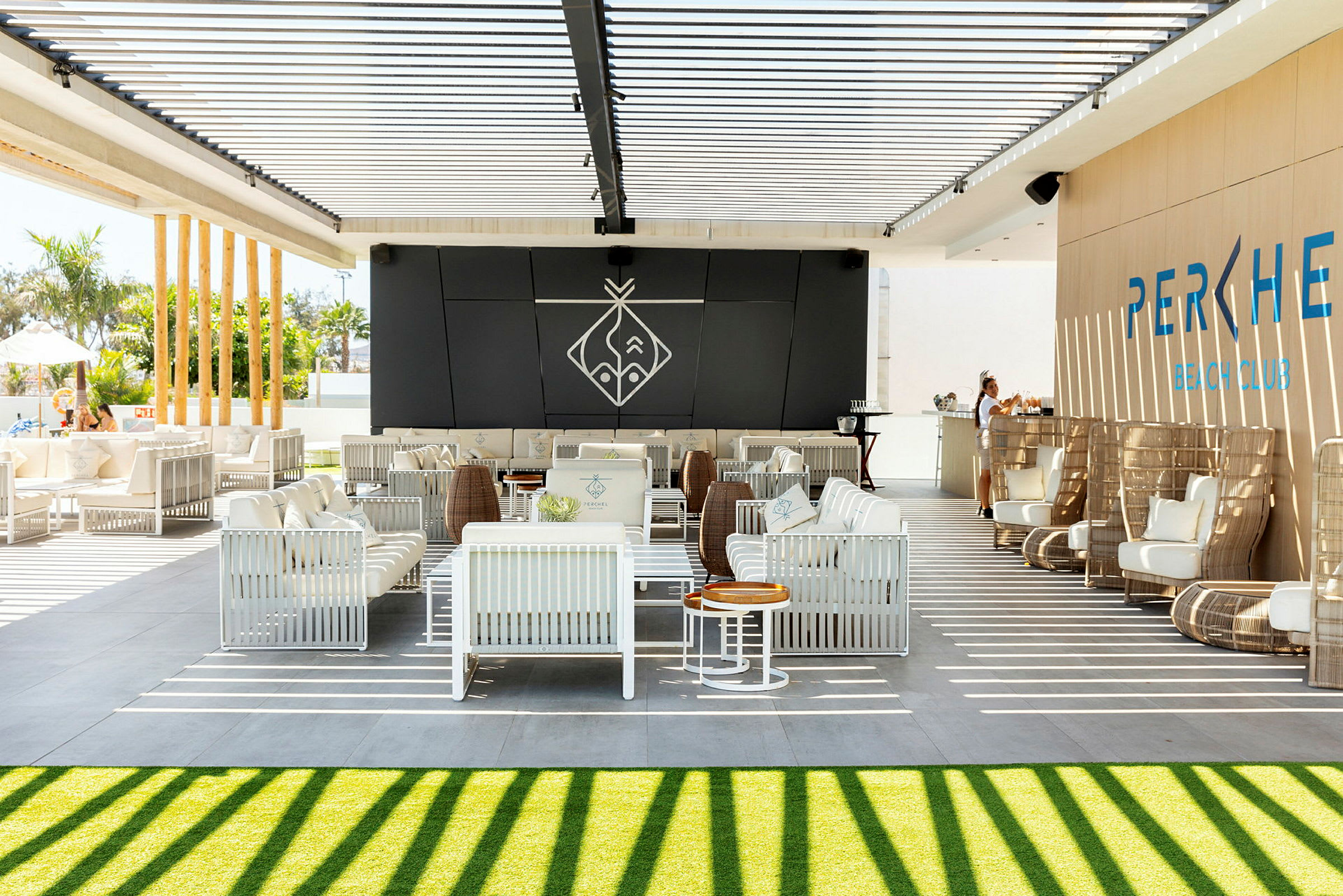 En udendørs lounge med hvide siddepladser og en bar under en pergola med skygger. (AI-genereret tekst)
