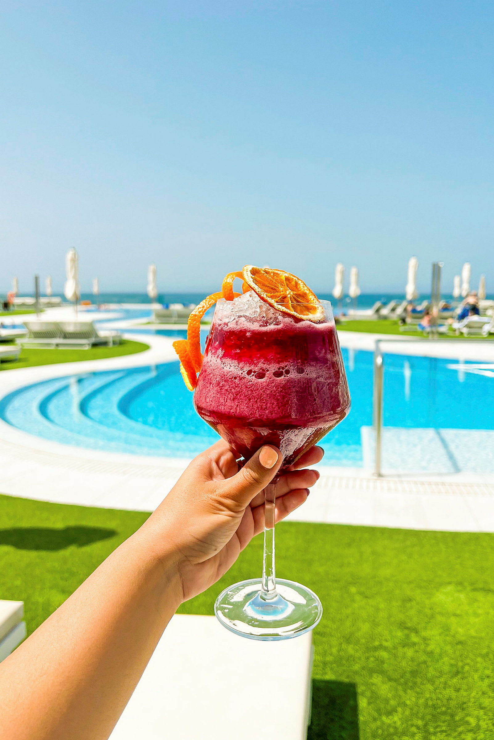 Et glas med rød drikk dekoreret med tørrede appelsinskiver holdes op foran en udendørs swimmingpool med liggestole og et blåt hav i baggrunden. (AI-genereret tekst)