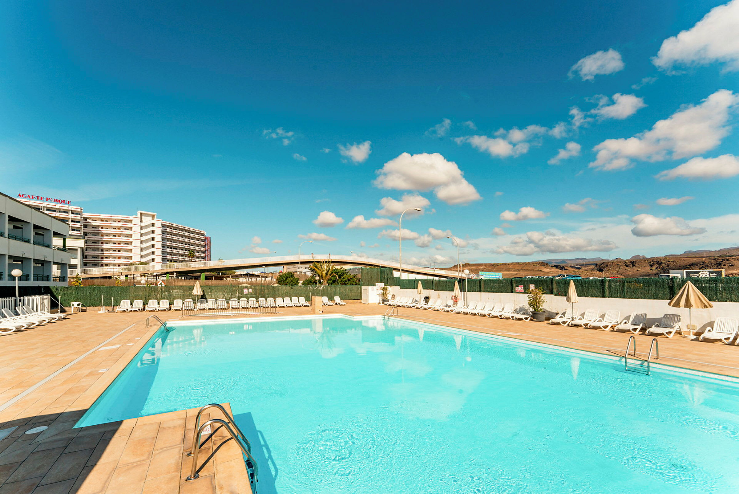 En udendørs pool omgivet af liggestole og parasoller med et hotel og en bro i baggrunden. (AI-genereret tekst)