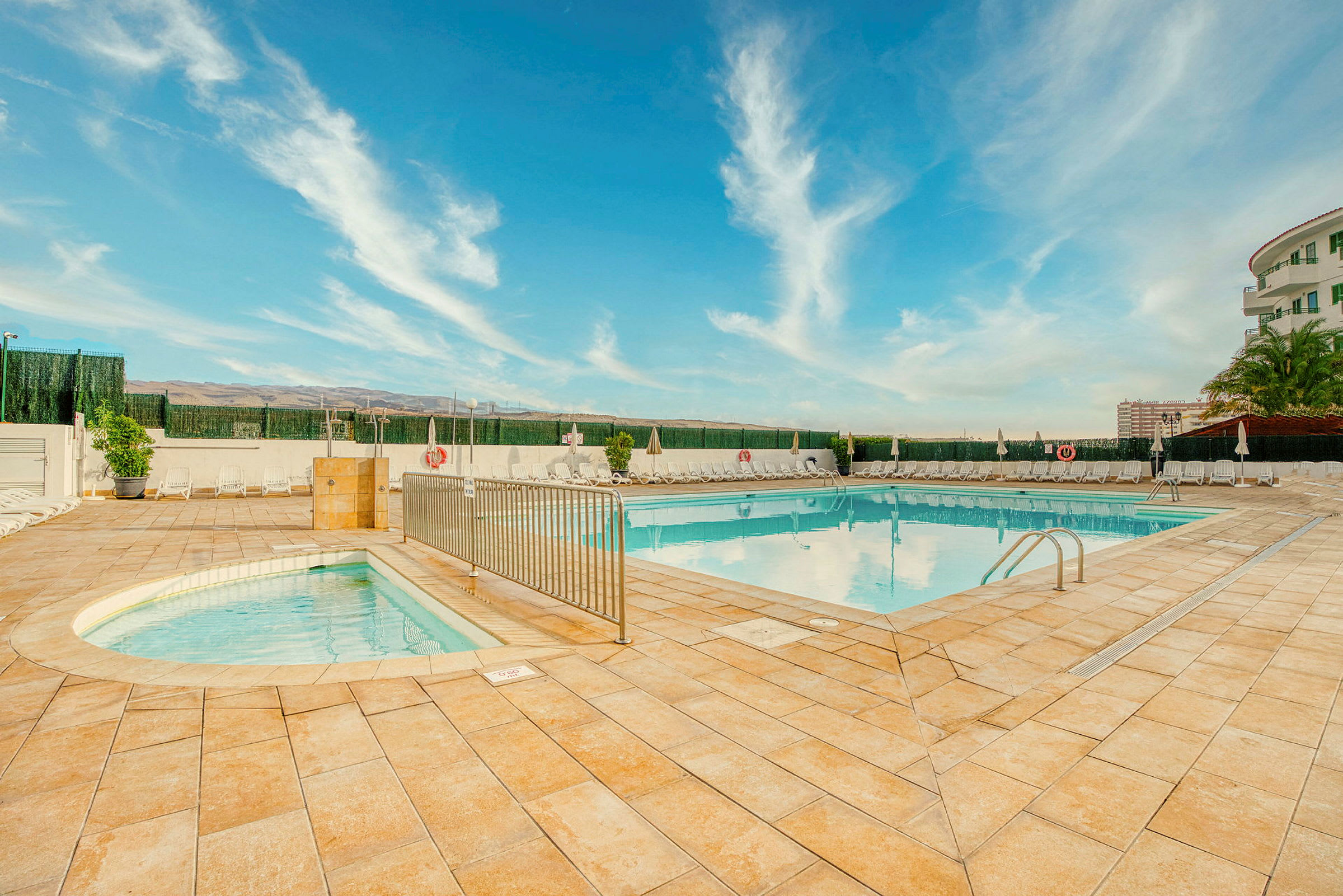 Et poolområde med to swimmingpools, omgivet af liggestole og parasoller, med en blå himmel i baggrunden. (AI-genereret tekst)