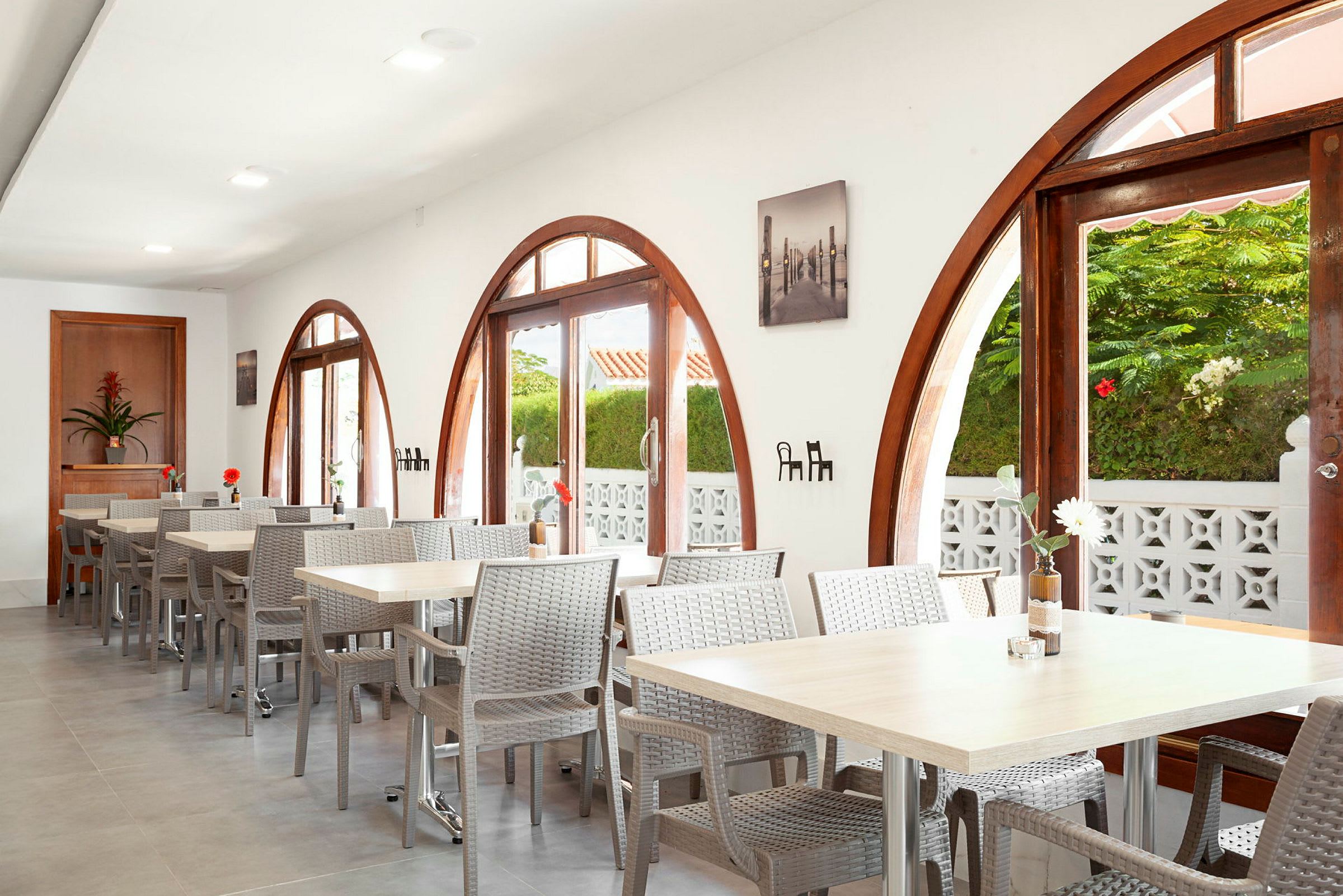 Restaurant med buede vinduer og borde med stole. Planter udenfor ses gennem vinduerne. (AI-genereret tekst)