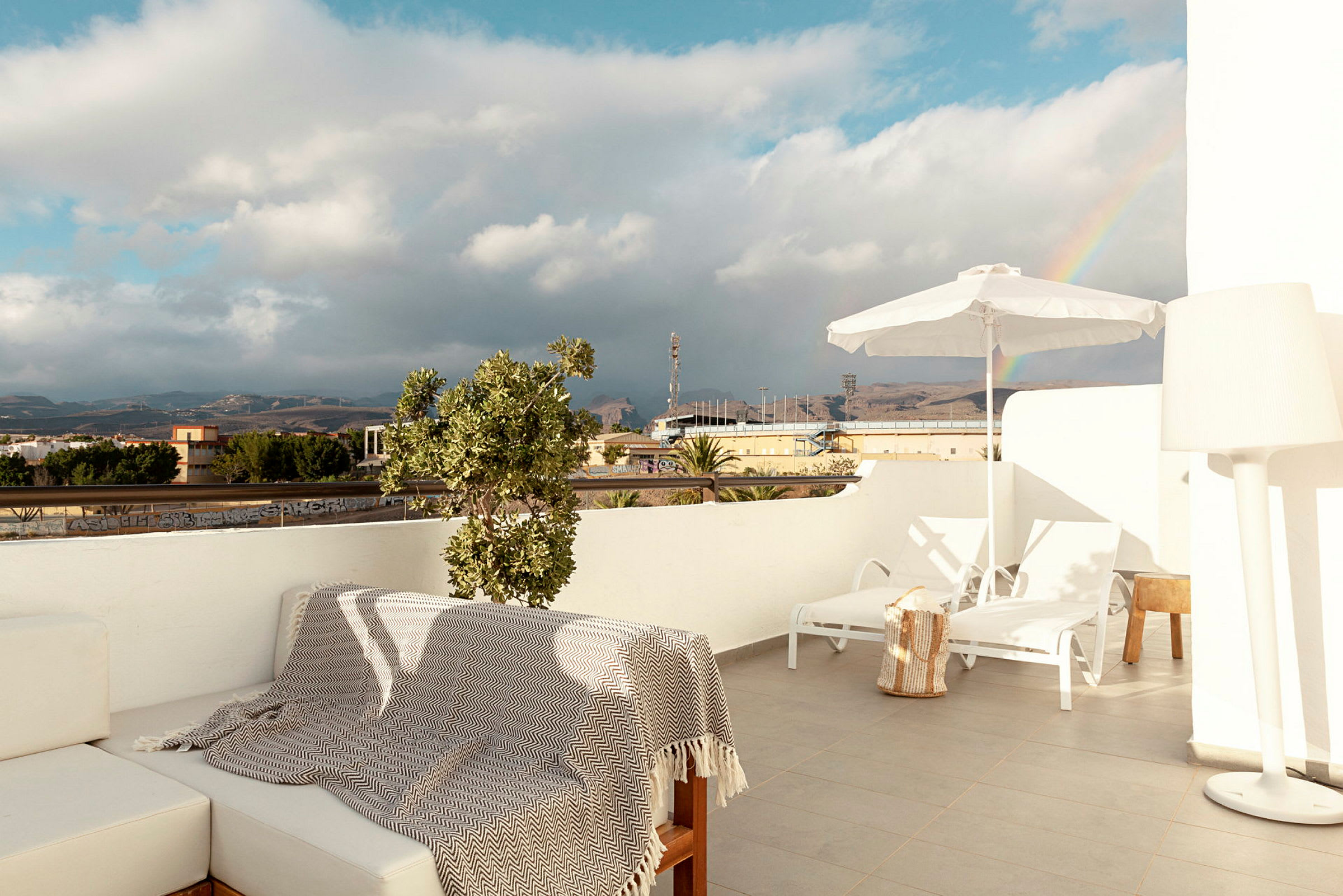 En terrasse med siddepladser, liggestole og parasol, med udsigt over et landskab med en regnbue i baggrunden. (AI-genereret tekst)