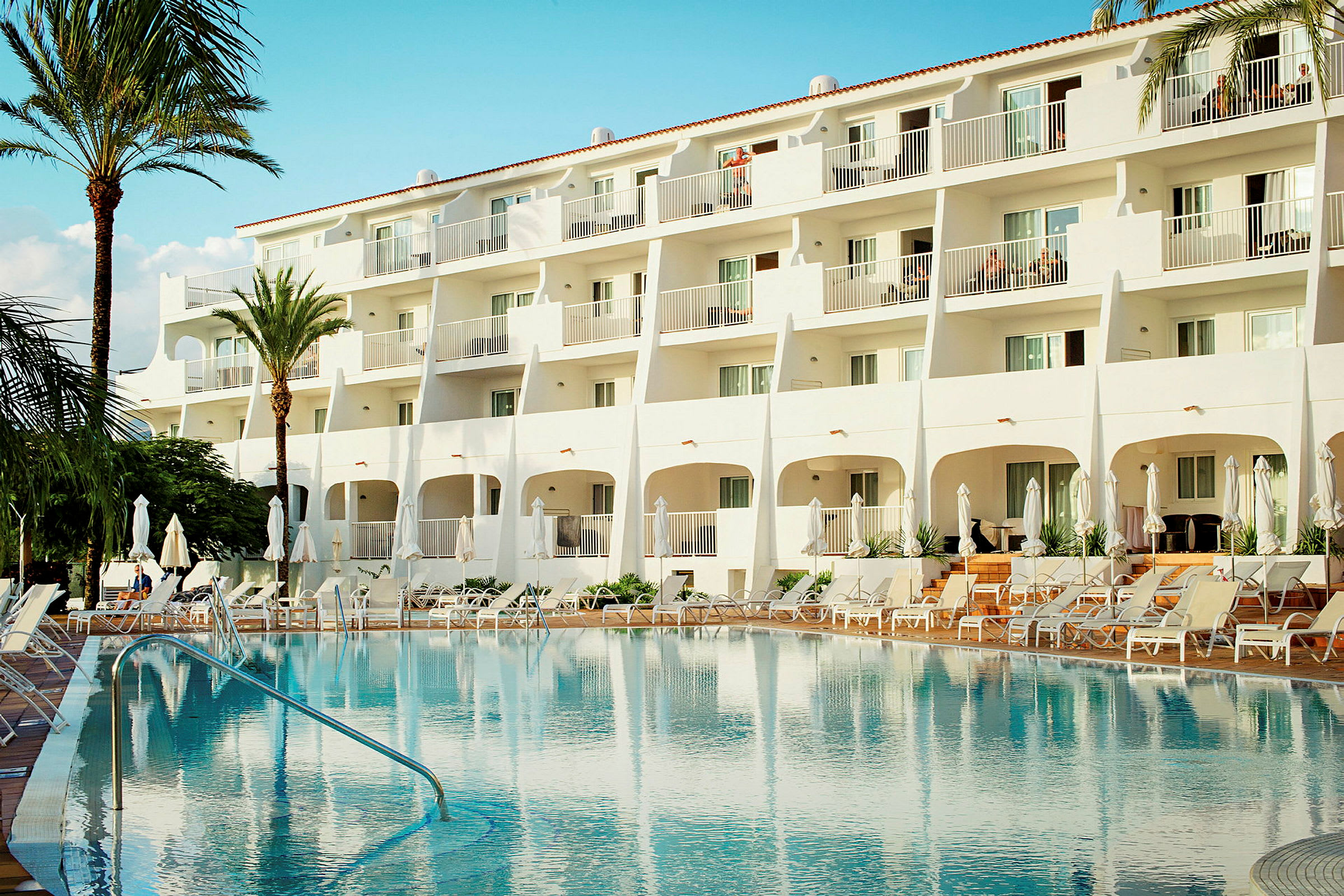Hotel med hvid facade og udendørs pool omgivet af liggestole. (AI-genereret tekst)