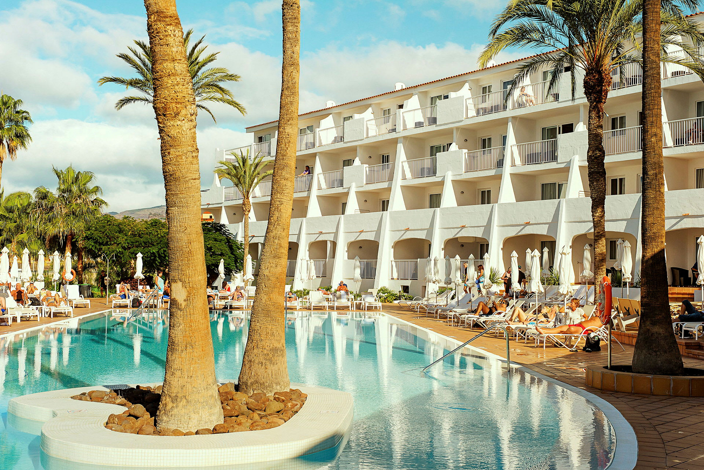 Hotelbygning ved en pool, omgivet af liggestole og palmer. (AI-genereret tekst)