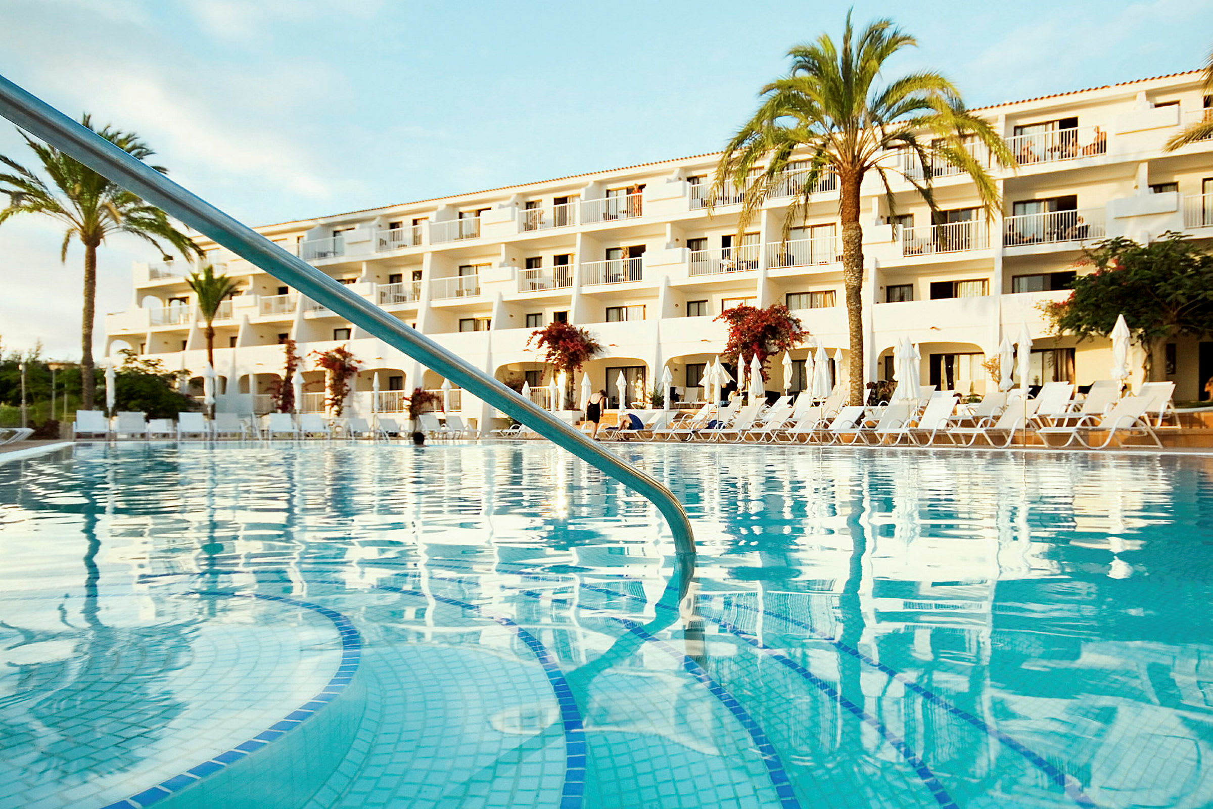 En udendørs swimmingpool foran en hvid bygning med altaner og palmetræer. (AI-genereret tekst)