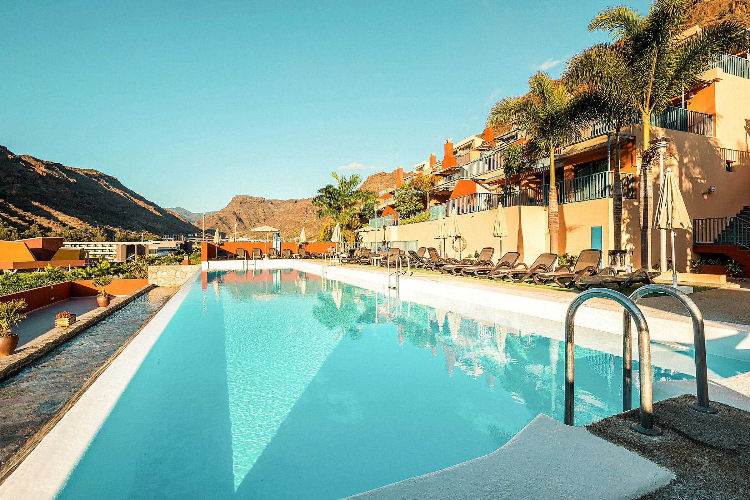 Udendørs pool ved et hotel med liggestole og bjerge i baggrunden. (AI-genereret tekst)