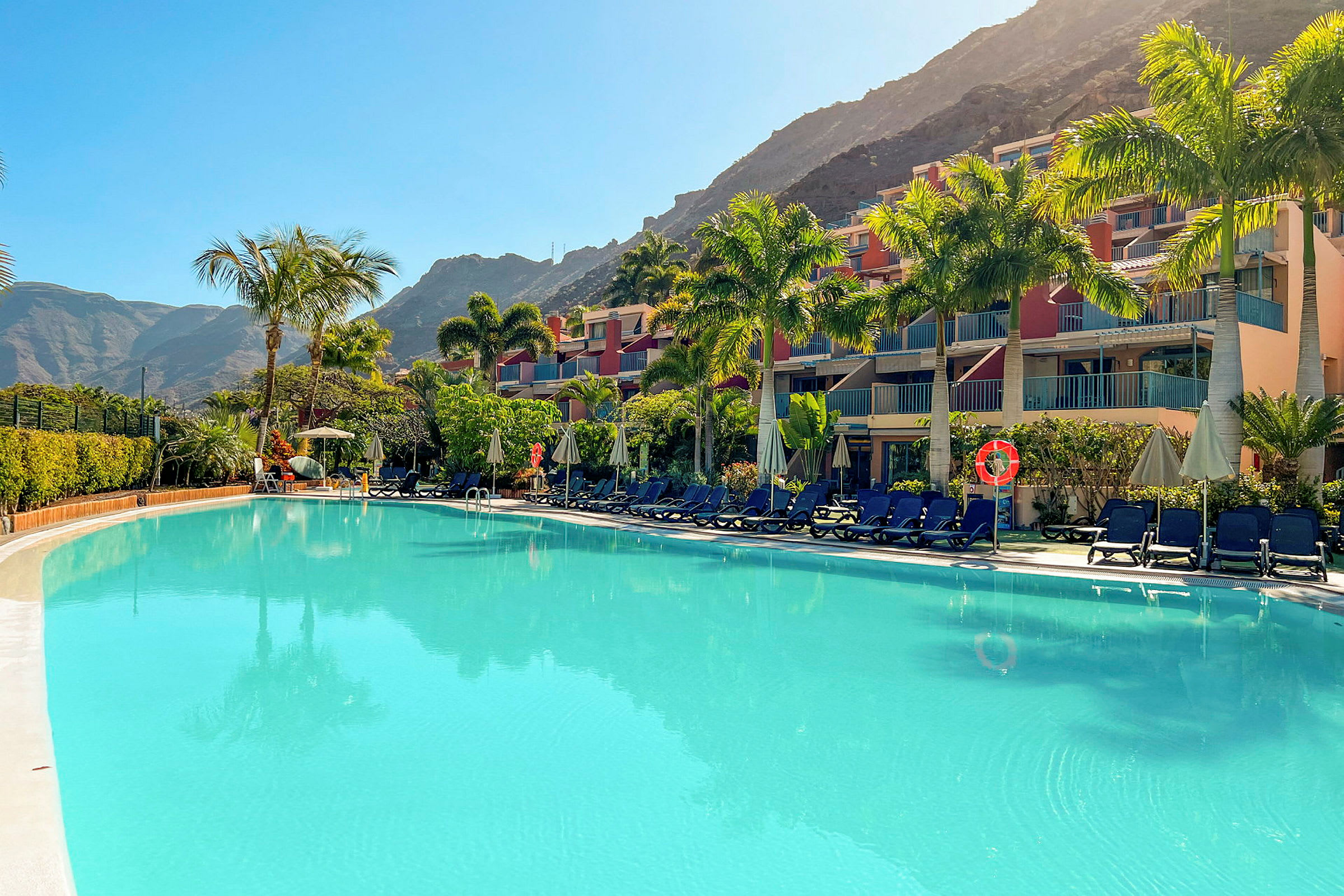 Billede af en swimmingpool, solsenge og hotelbygninger omgivet af palmer ved foden af et bjerg. (AI-genereret tekst)