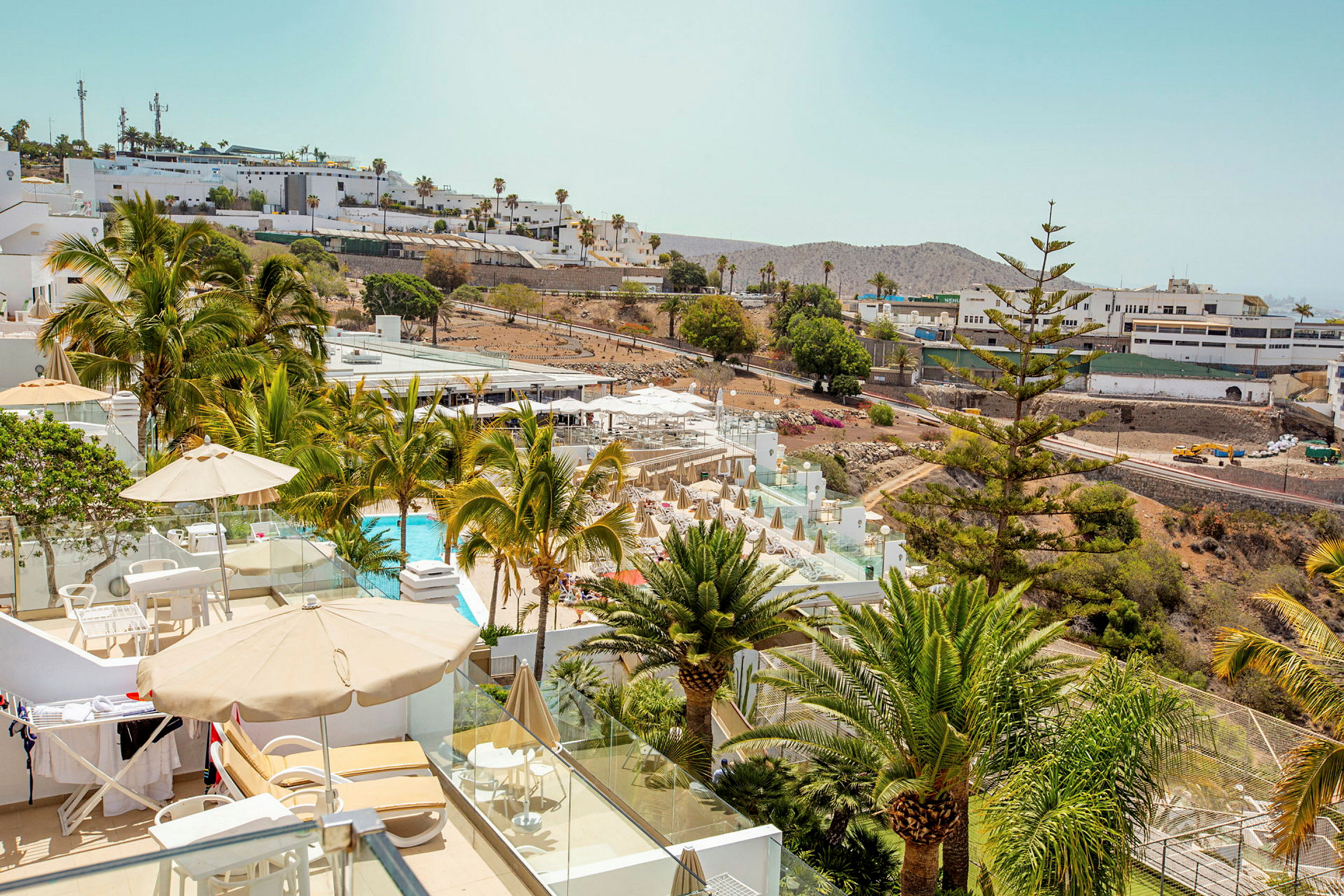 Udsigt over en hotelpool og terrasse med liggestole og parasoller, omgivet af grønt og palmer. (AI-genereret tekst)