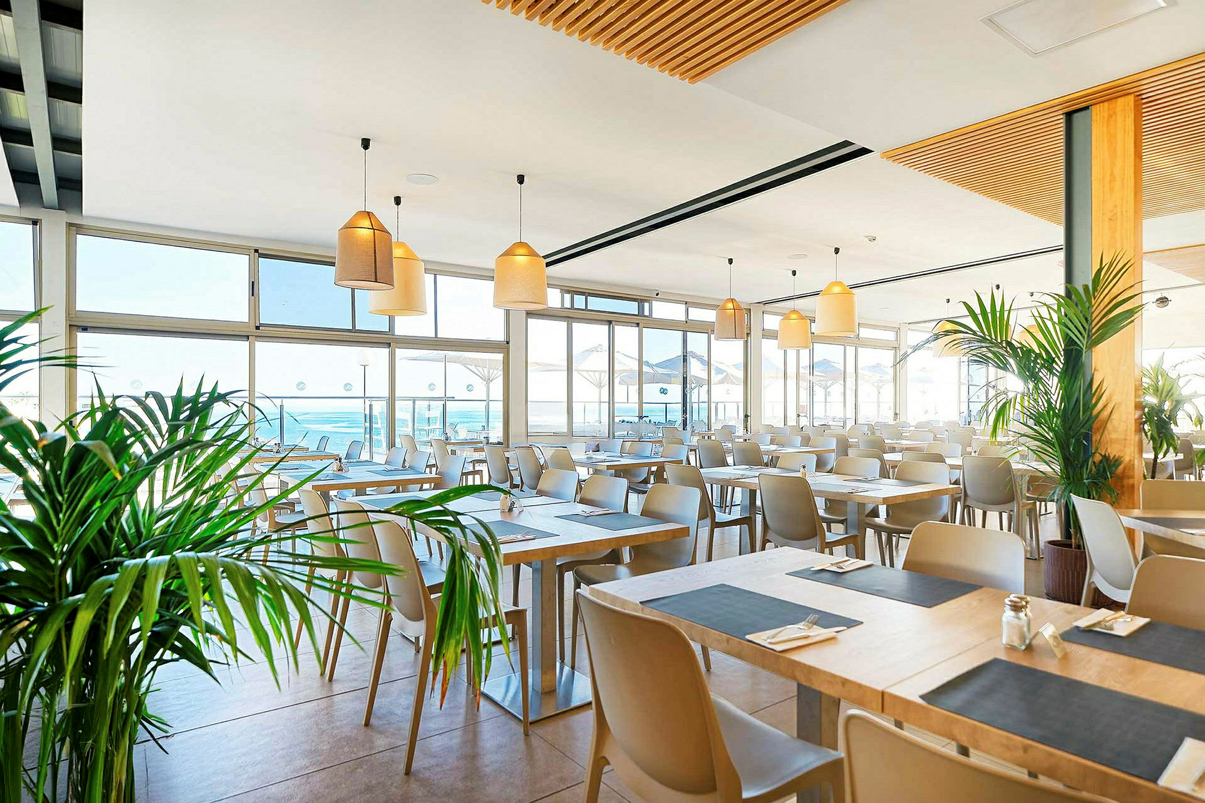 Lys og moderne restaurant med mange borde, store vinduer og planter. (AI-genereret tekst)