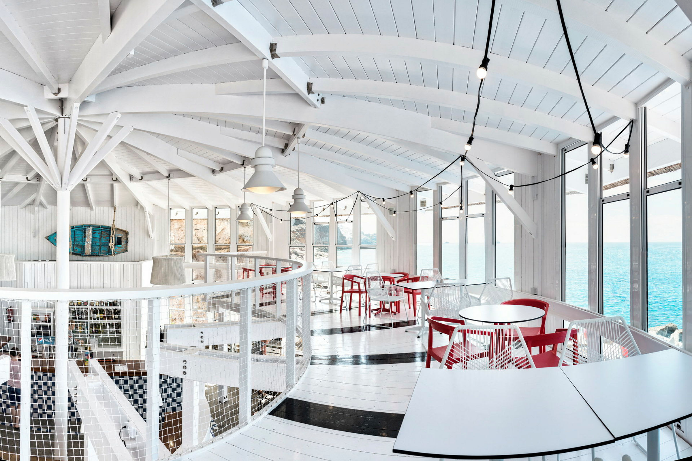 Lyst indrettet restaurant med hvide vægge, røde stole og store vinduer med udsigt over havet. (AI-genereret tekst)