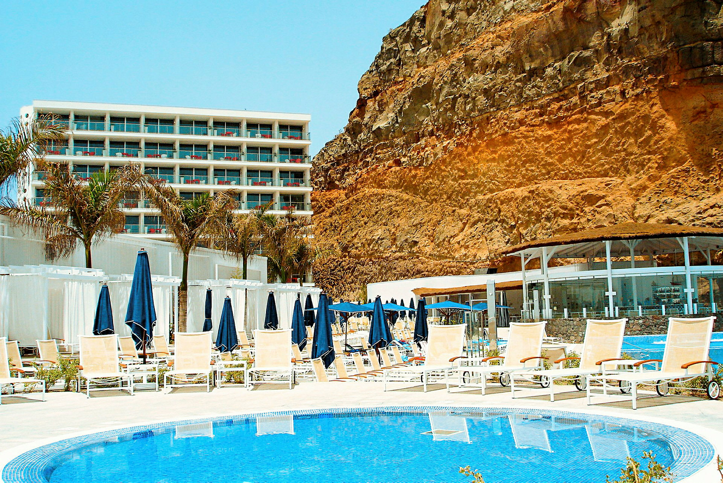 Billede af en hotelpool med liggestole og parasoller, med et hotelkompleks i baggrunden og en stor klippevæg ved siden af. (AI-genereret tekst)