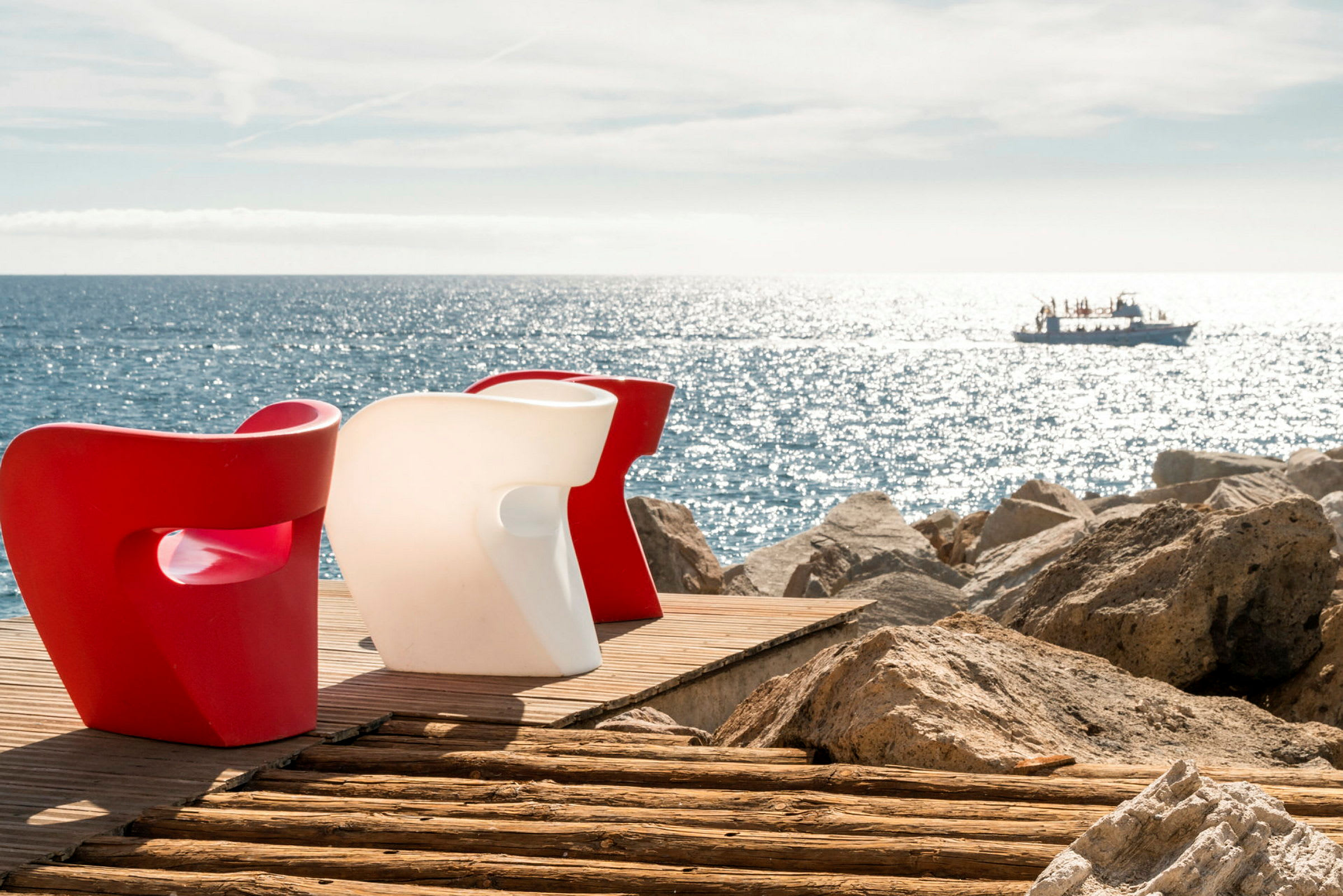 To moderne stole, en rød og en hvid, står på en træterrasse ved havet med en båd synlig i baggrunden. (AI-genereret tekst)