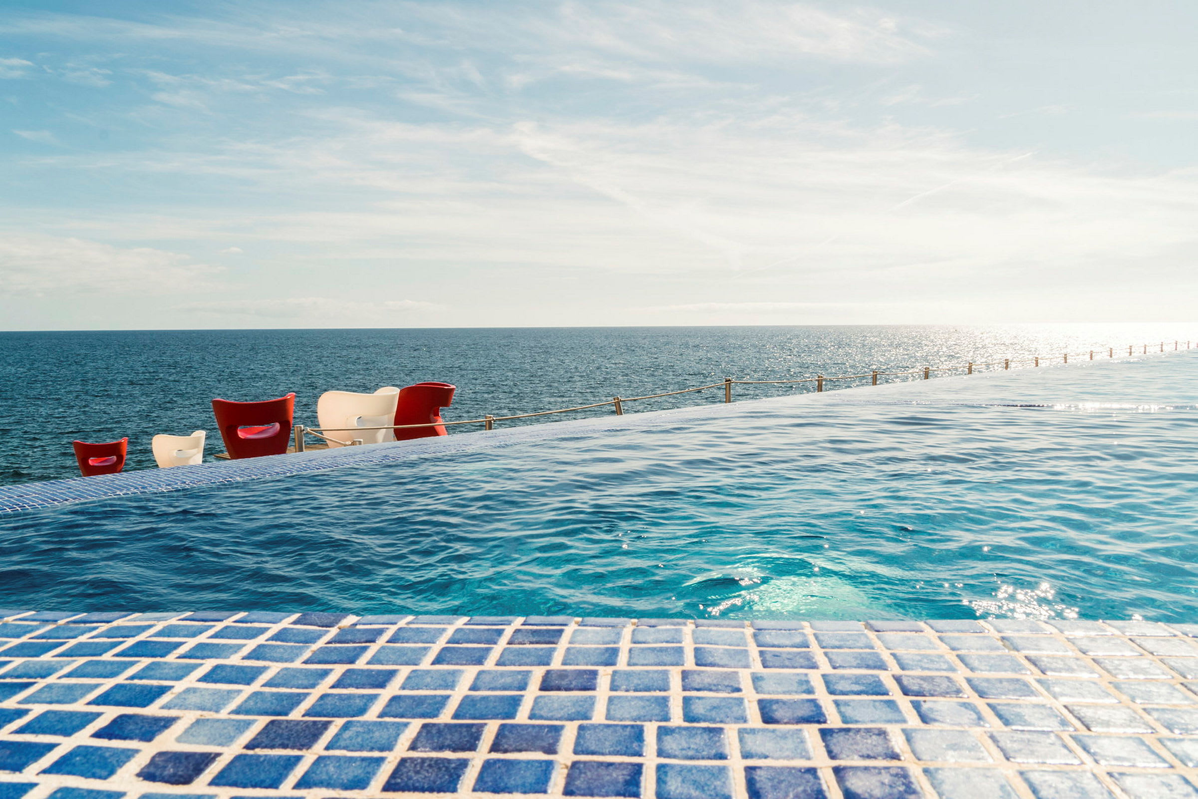 Udsigt over en infinitypool med blå mosaik, der strækker sig mod havet og himlen. (AI-genereret tekst)