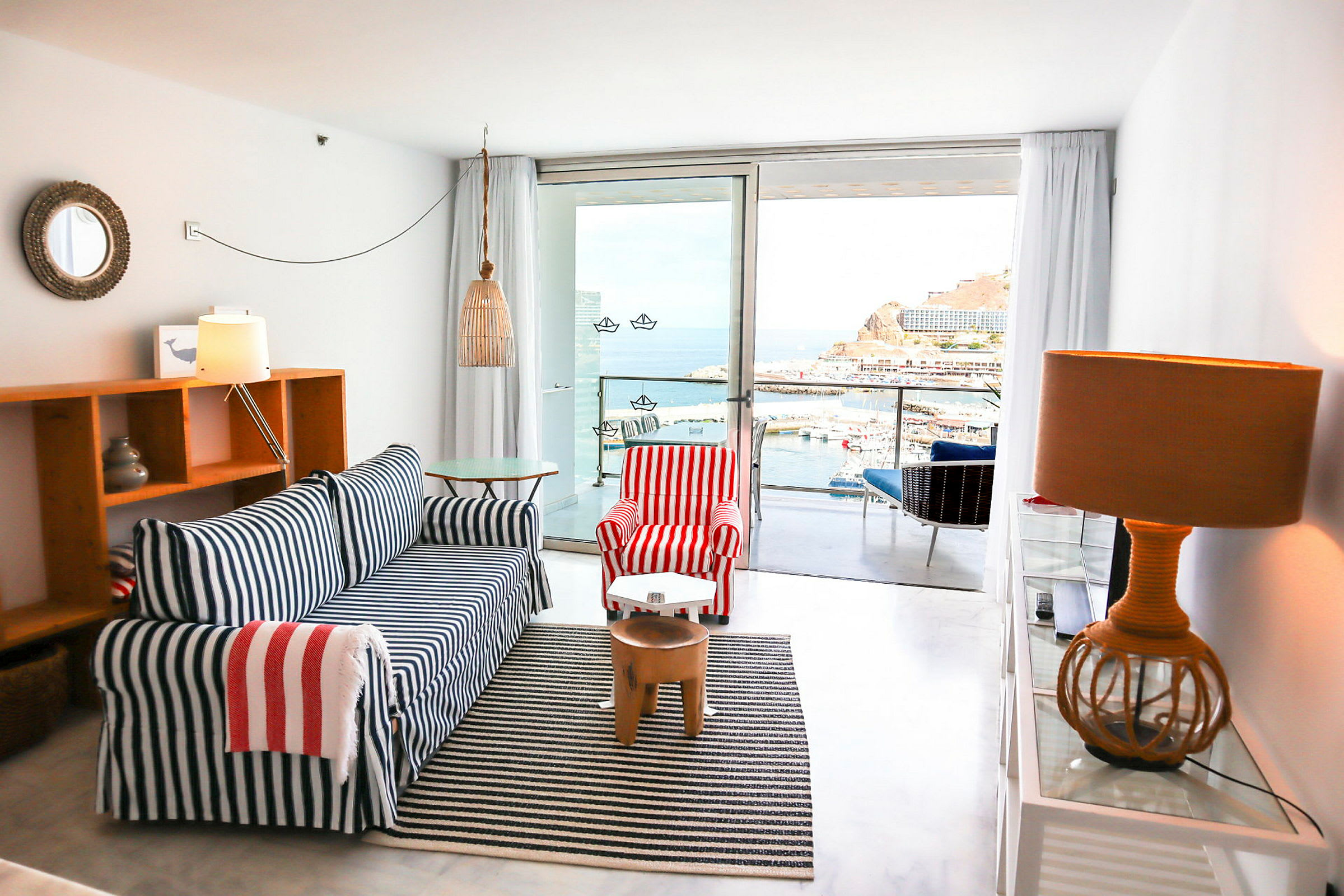 Et hotelværelse med maritim indretning, sofaen har blå og hvide striber, lænestolen har røde og hvide striber. Udsigt over en havn og hav fra en altan. (AI-genereret tekst)