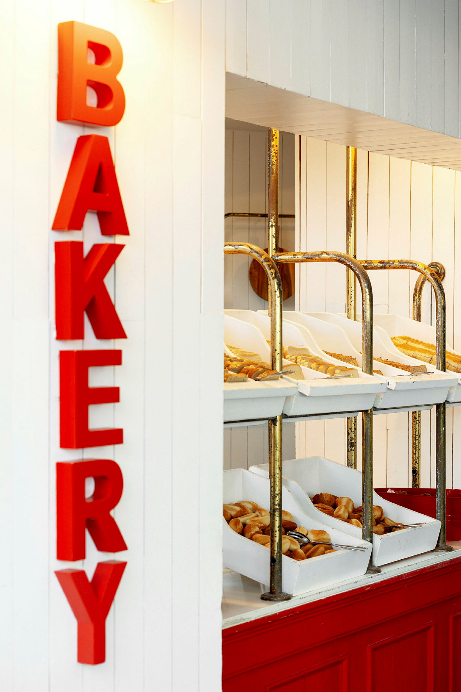 Et bageriområde med røde bogstaver, der staver ordet 'BAKERY'. Hylder indeholder forskellige typer brød og bagværk i hvide kasser. (AI-genereret tekst)