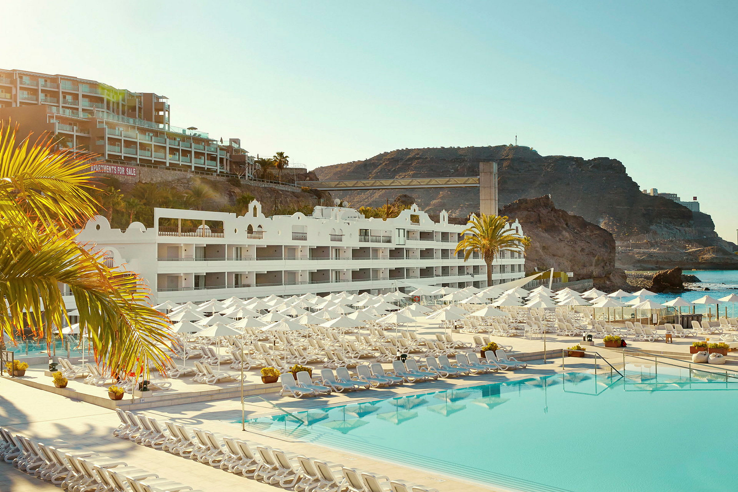 Hotelbygning med hvide facader, liggestole og parasoller ved en stor pool, samt klippeformationer i baggrunden. (AI-genereret tekst)