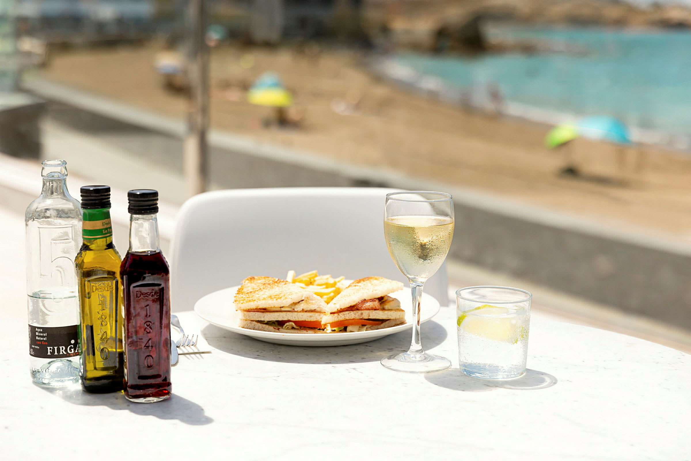 En tallerken med sandwich og pommes frites, omgivet af flasker, et glas hvidvin og et glas vand med lime, med en strand i baggrunden. (AI-genereret tekst)