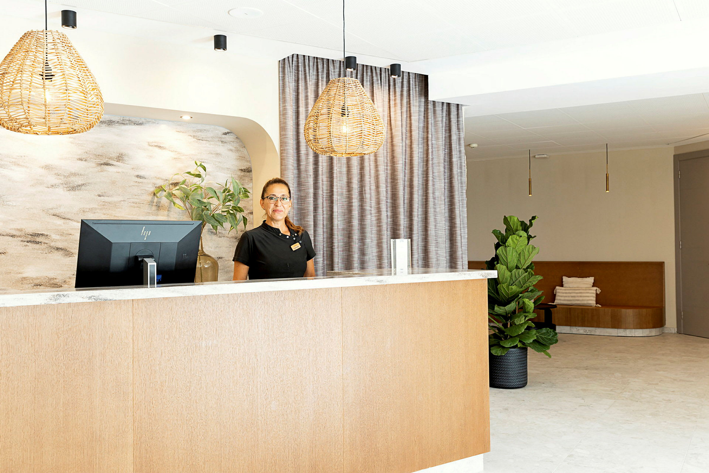 Reception med skranke og computer, vægdekorationer og indretningsdetaljer som gardiner og grøn plante. (AI-genereret tekst)