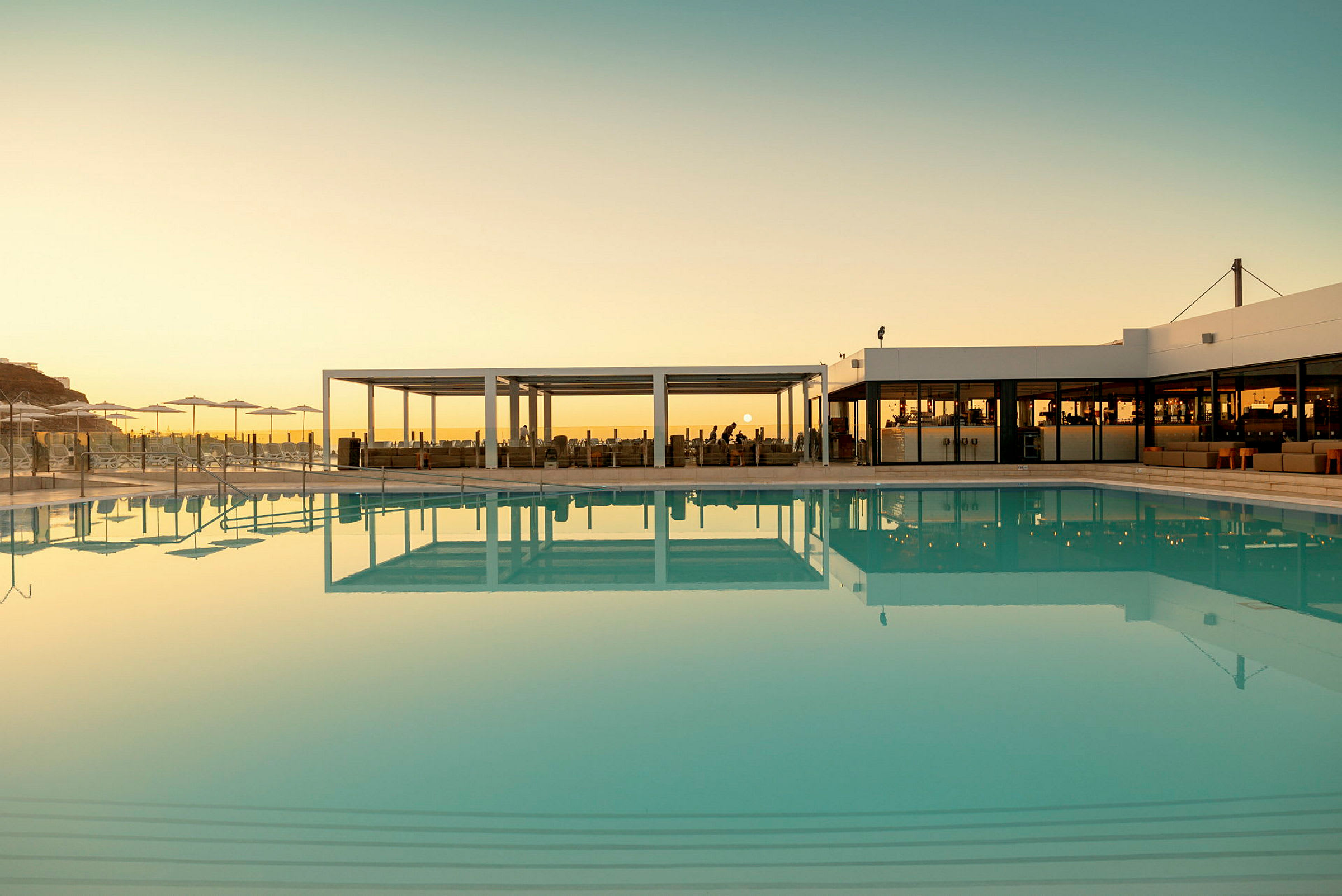 En udendørs pool med klart vand ved solnedgang, omgivet af bygninger og parasoller. (AI-genereret tekst)