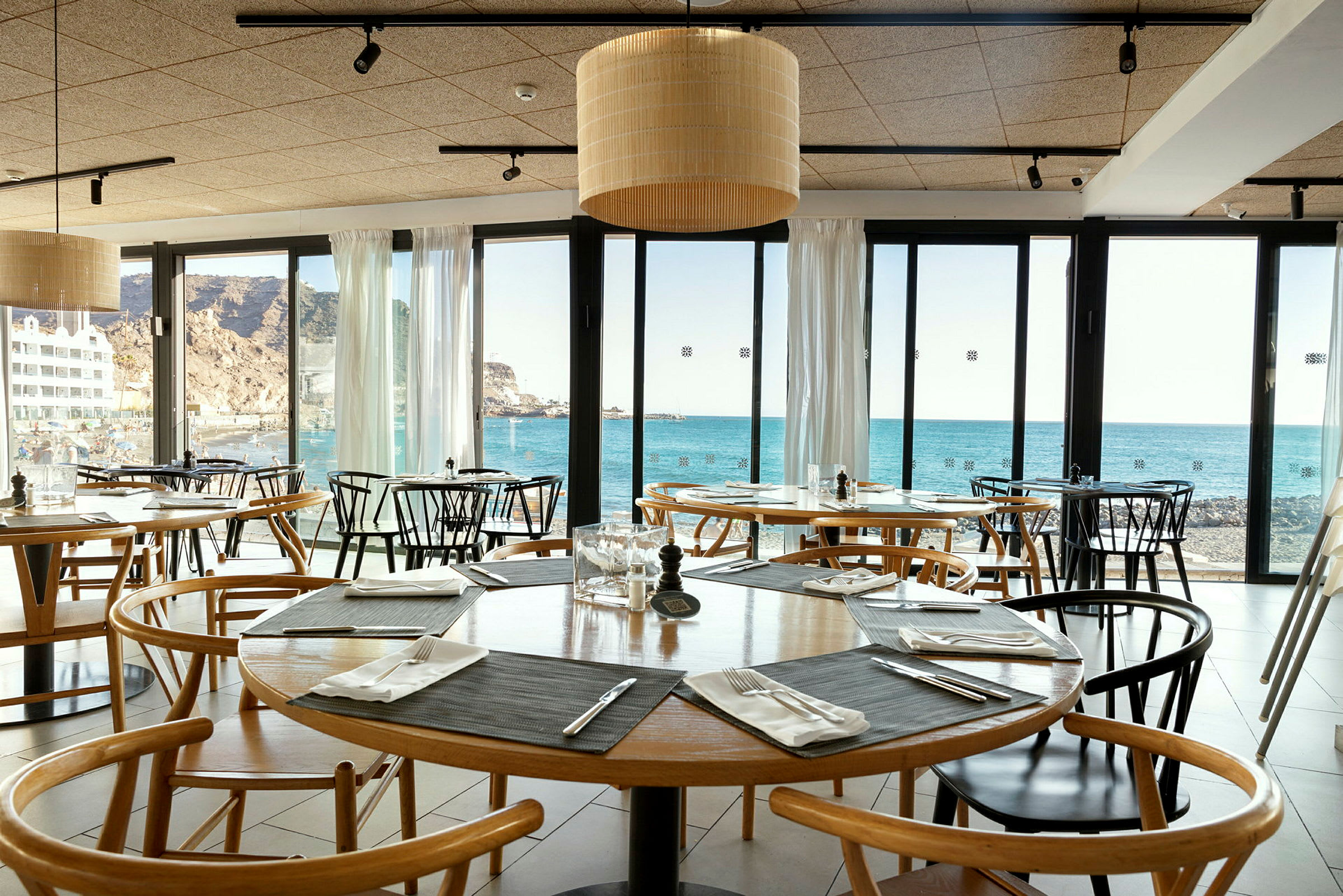 Indersiden af en restaurant med borde og stole, udsigt til havet gennem glasvinduer. (AI-genereret tekst)
