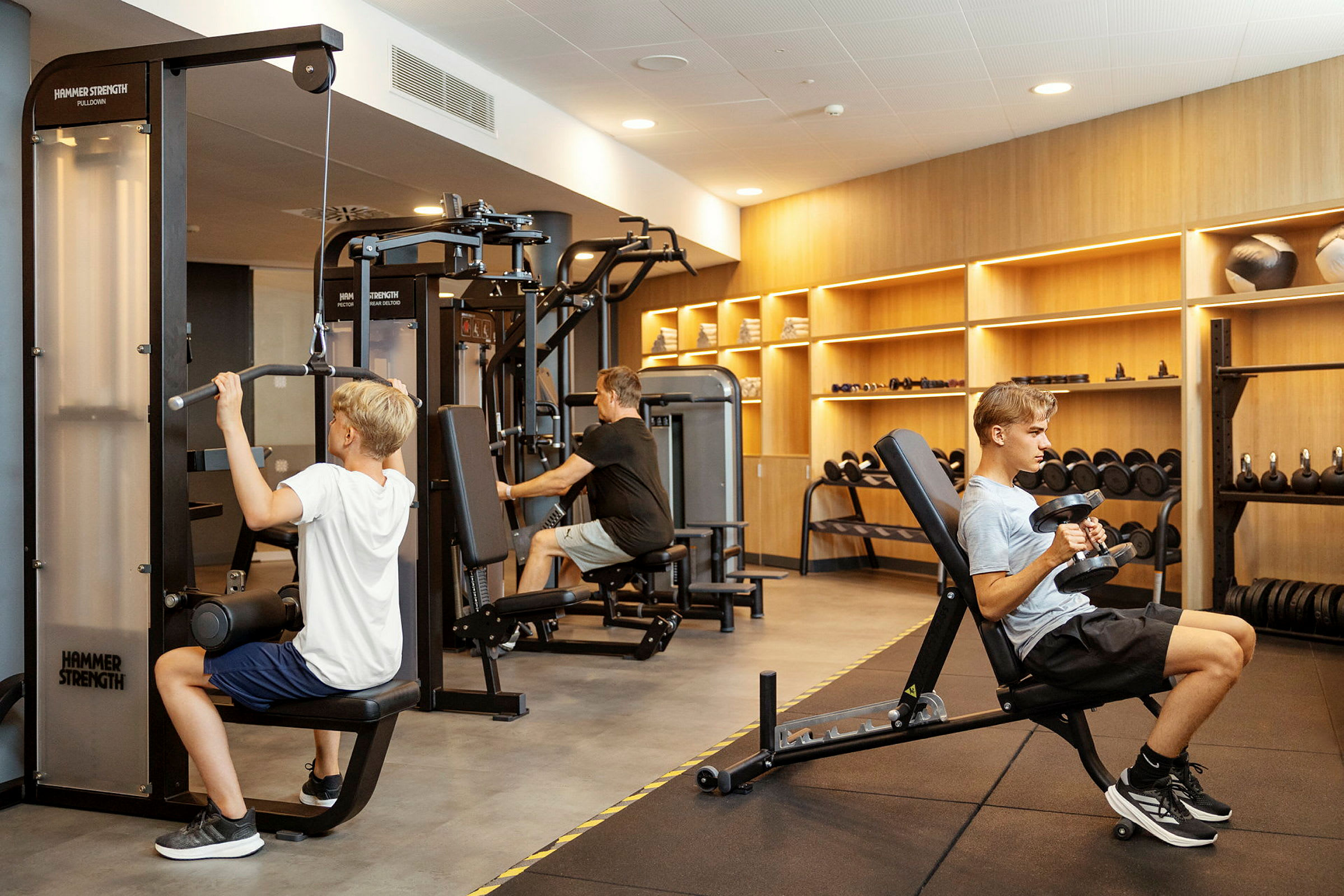 Personer der træner i et fitnesscenter med forskellige træningsmaskiner og vægte. (AI-genereret tekst)