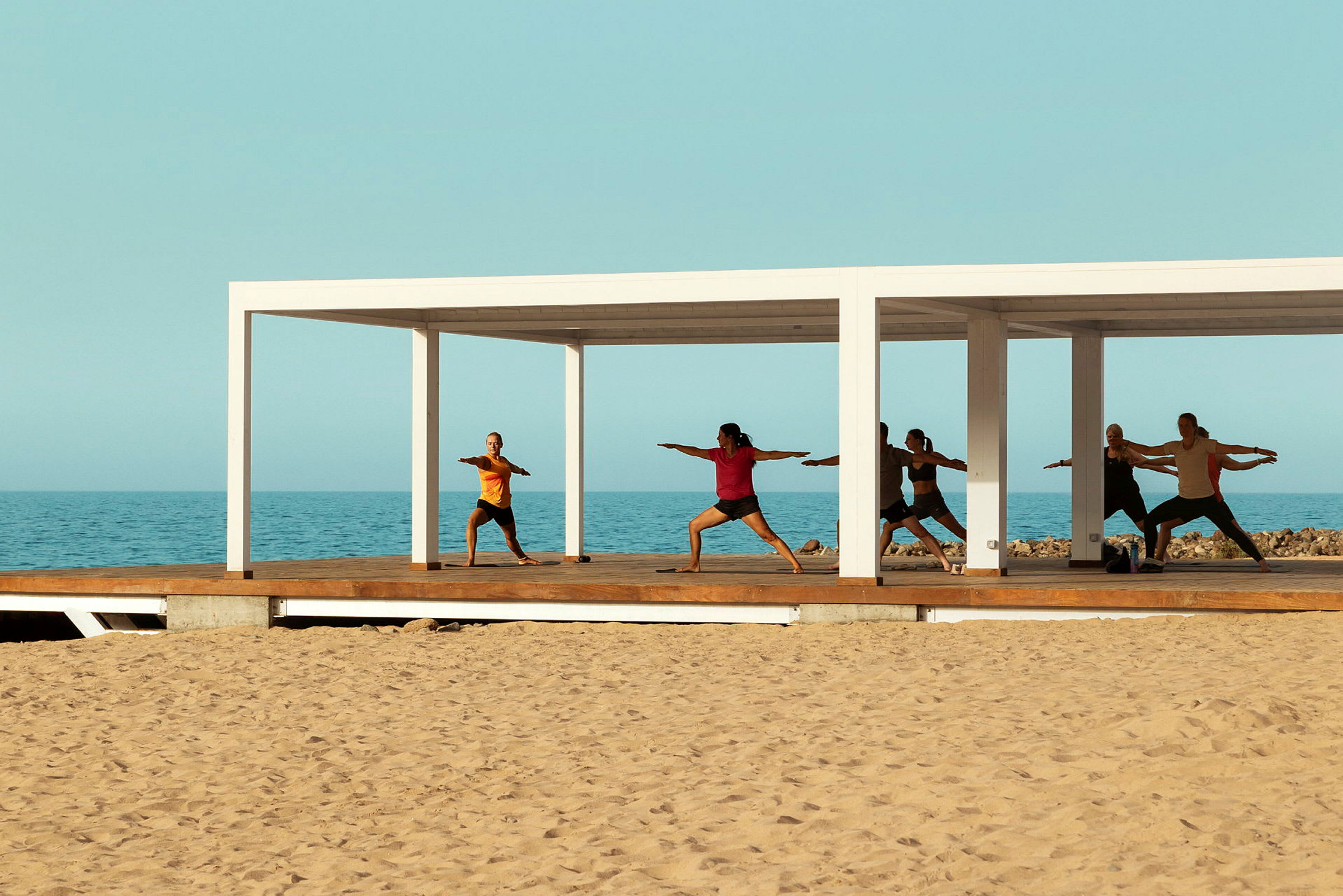 Personer, der laver yoga på en træplatform ved havet under en hvid pergola. (AI-genereret tekst)