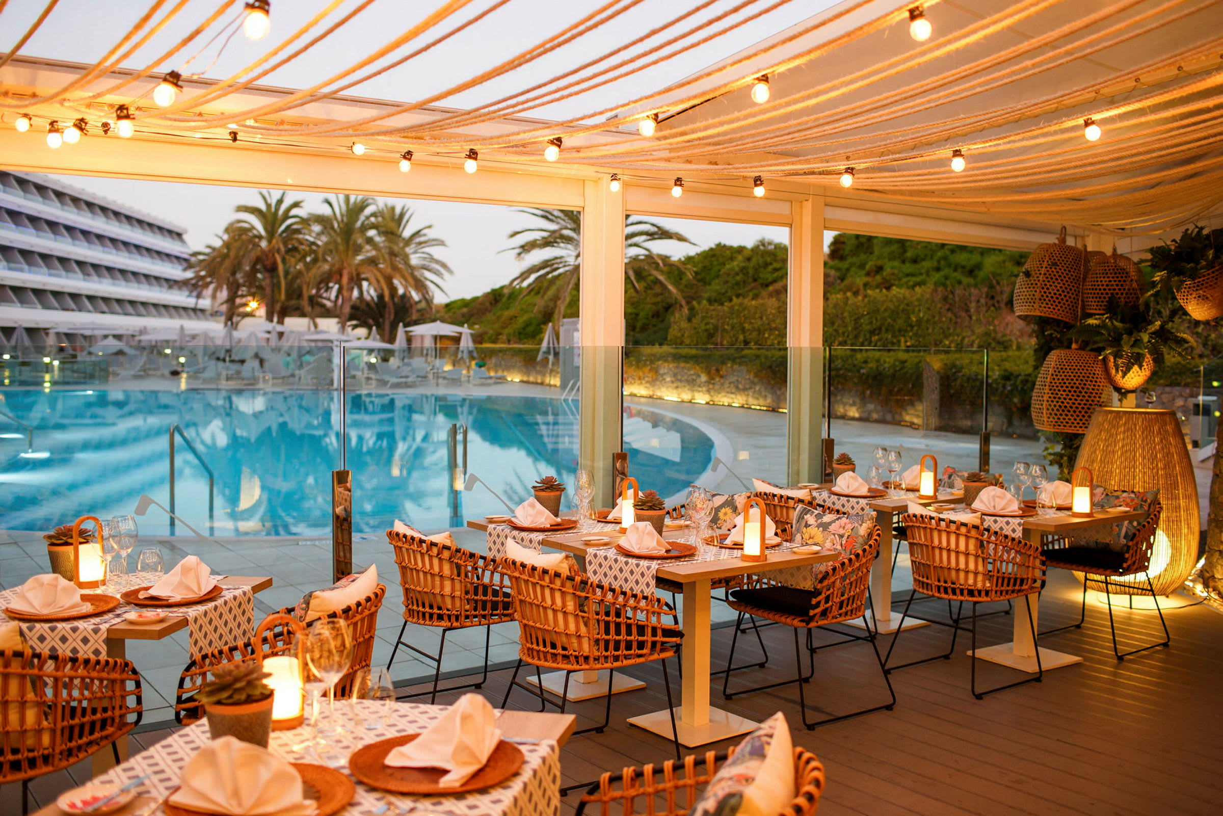 Udendørs restaurant med dækkede borde og lyskæder, beliggende tæt på en pool og omgivet af palmer og grøntområder. (AI-genereret tekst)