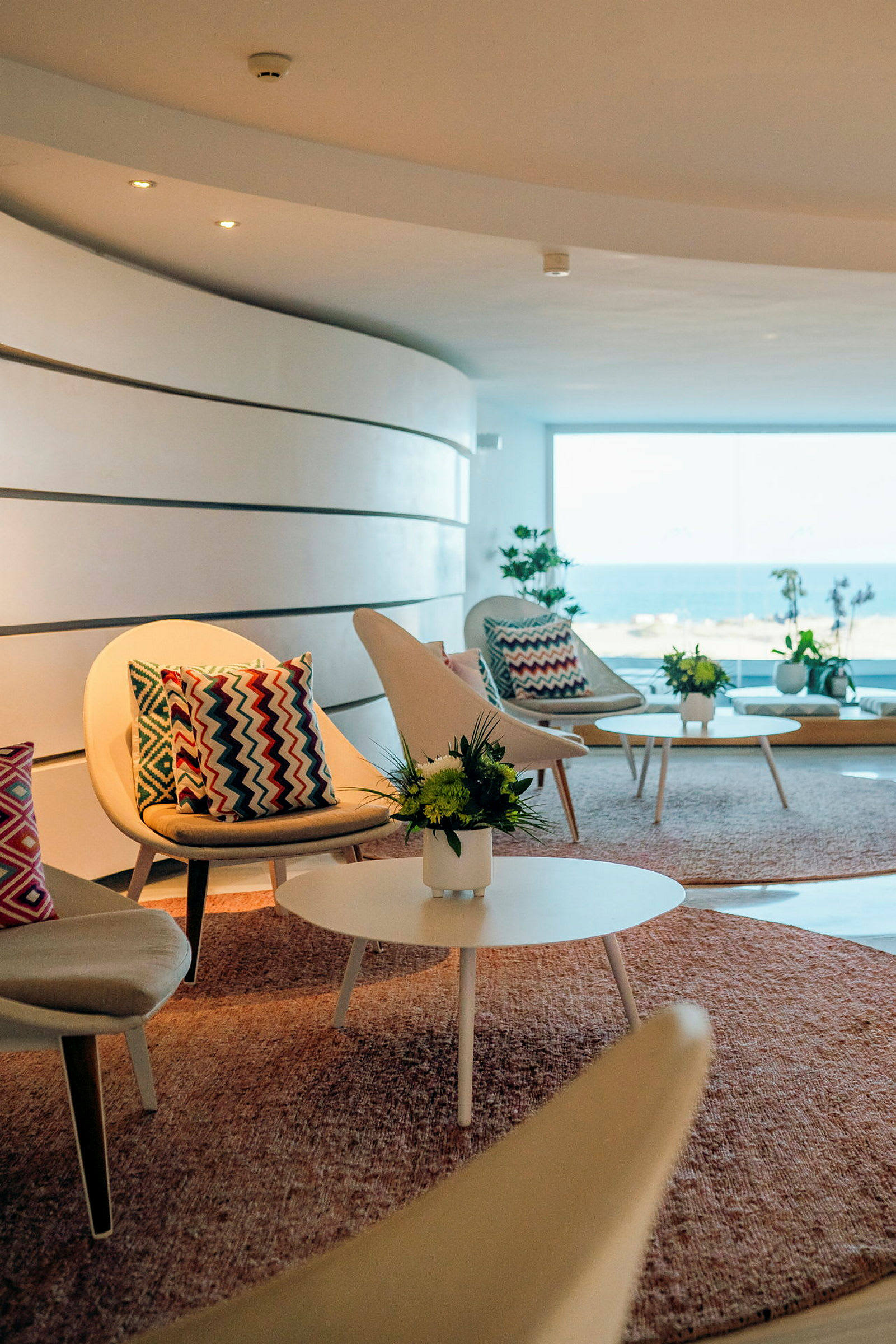 En lounge med moderne stole og sofaborde, dekoreret med farverige puder og blomster, med havudsigt gennem et stort vindue. (AI-genereret tekst)