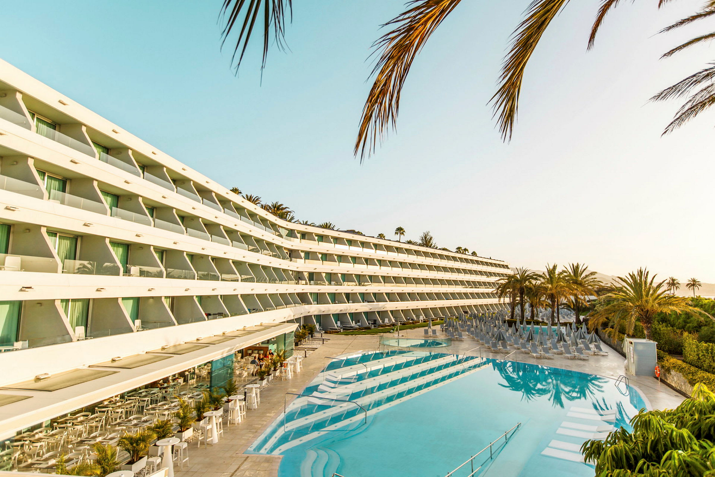 Moderne hotelbygning med udsigt over en solrig pool og omkringliggende palmer. (AI-genereret tekst)