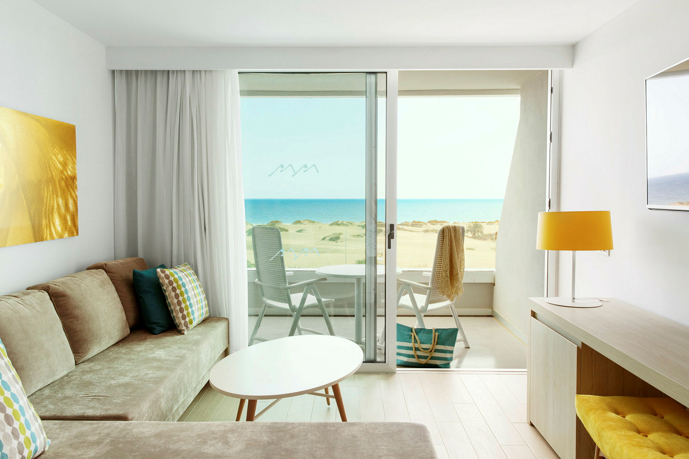 Udsigt fra et lyst hotelværelse med moderne indretning over en solrig strand og havet. (AI-genereret tekst)