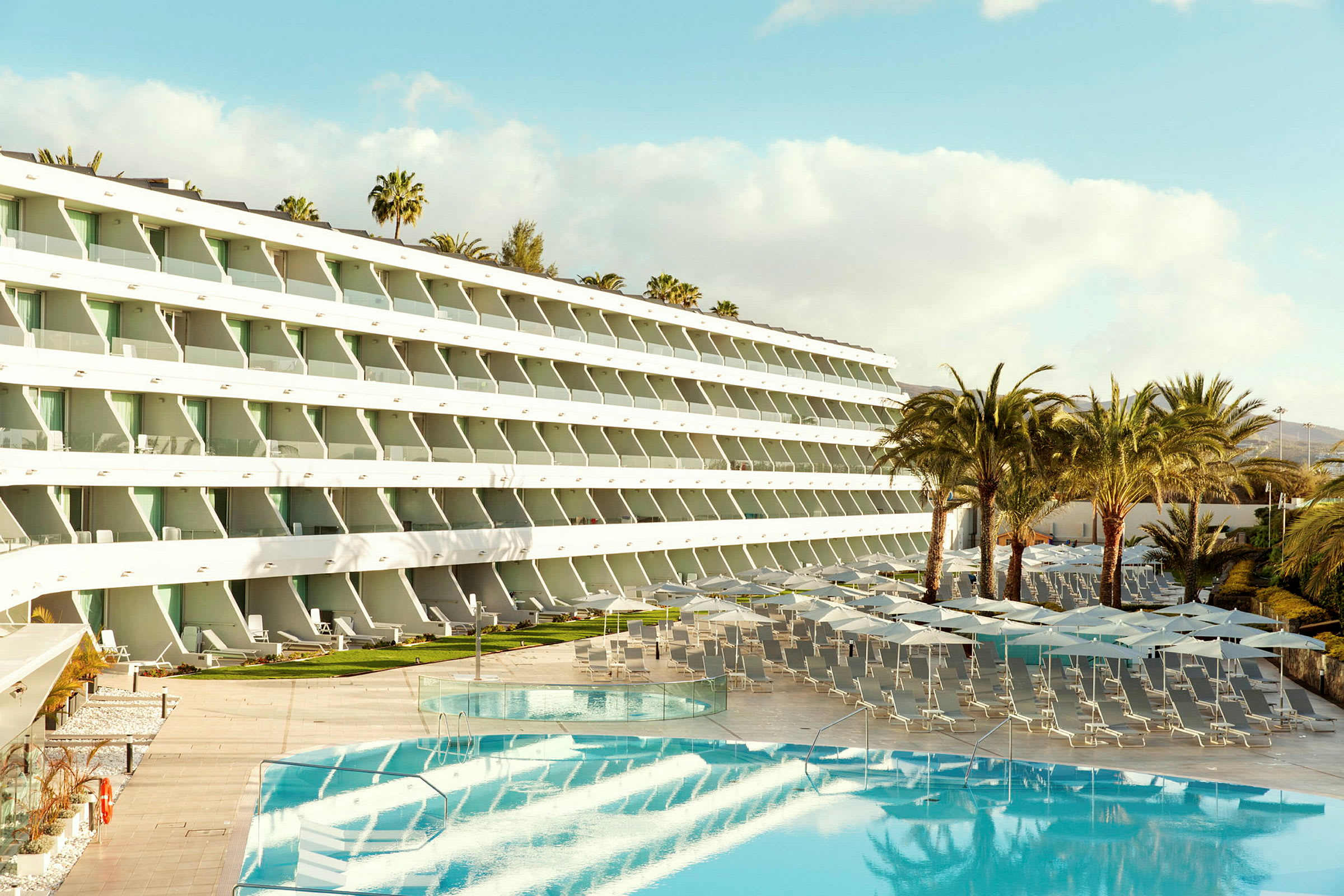 Hotelkompleks med pool, liggestole og parasoller, omgivet af palmer. (AI-genereret tekst)
