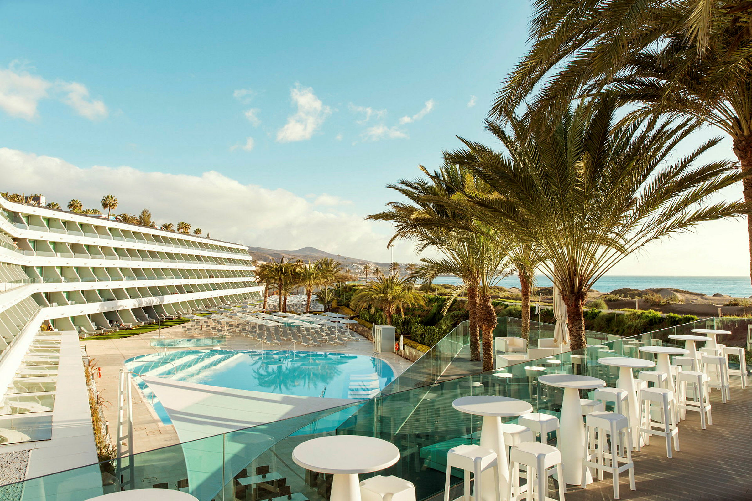 Billede af et hotel med udsigt over en pool og palmer, med havet i baggrunden. (AI-genereret tekst)