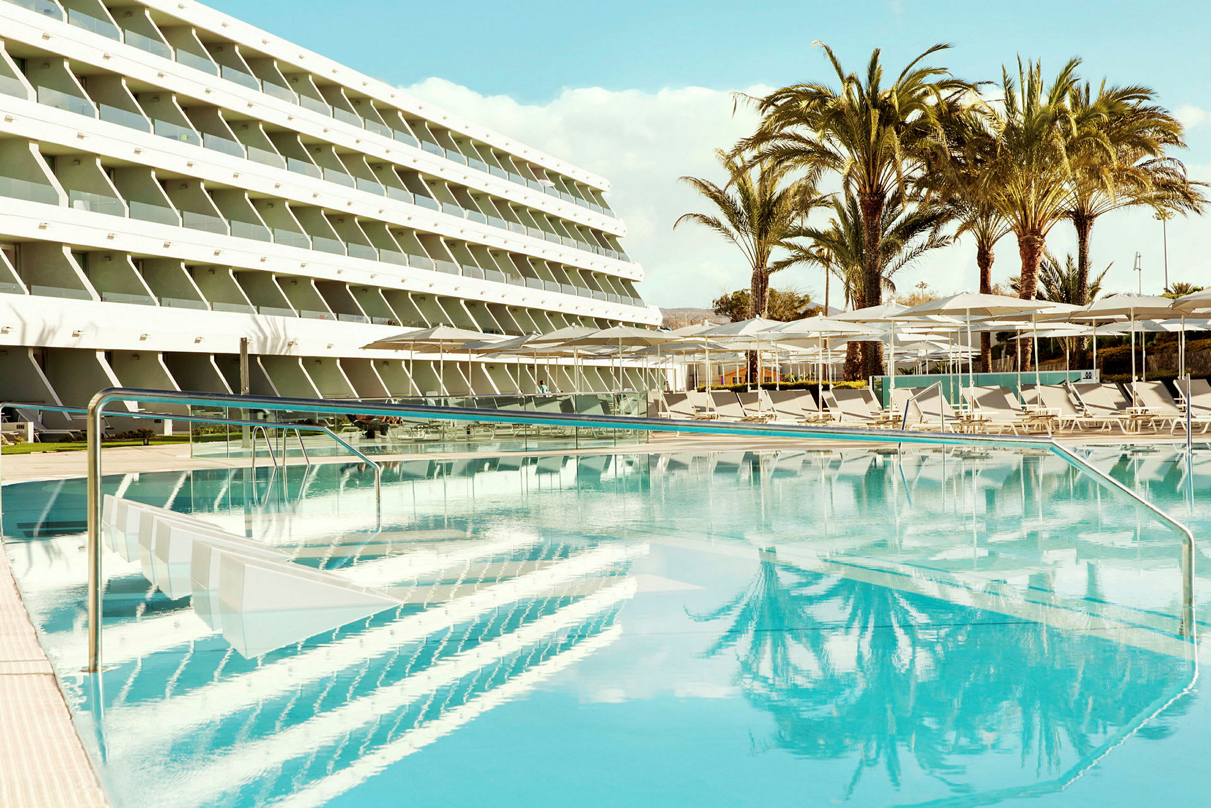 Poolområdet med liggestole, parasoller og et fleretagers hotel i baggrunden. (AI-genereret tekst)
