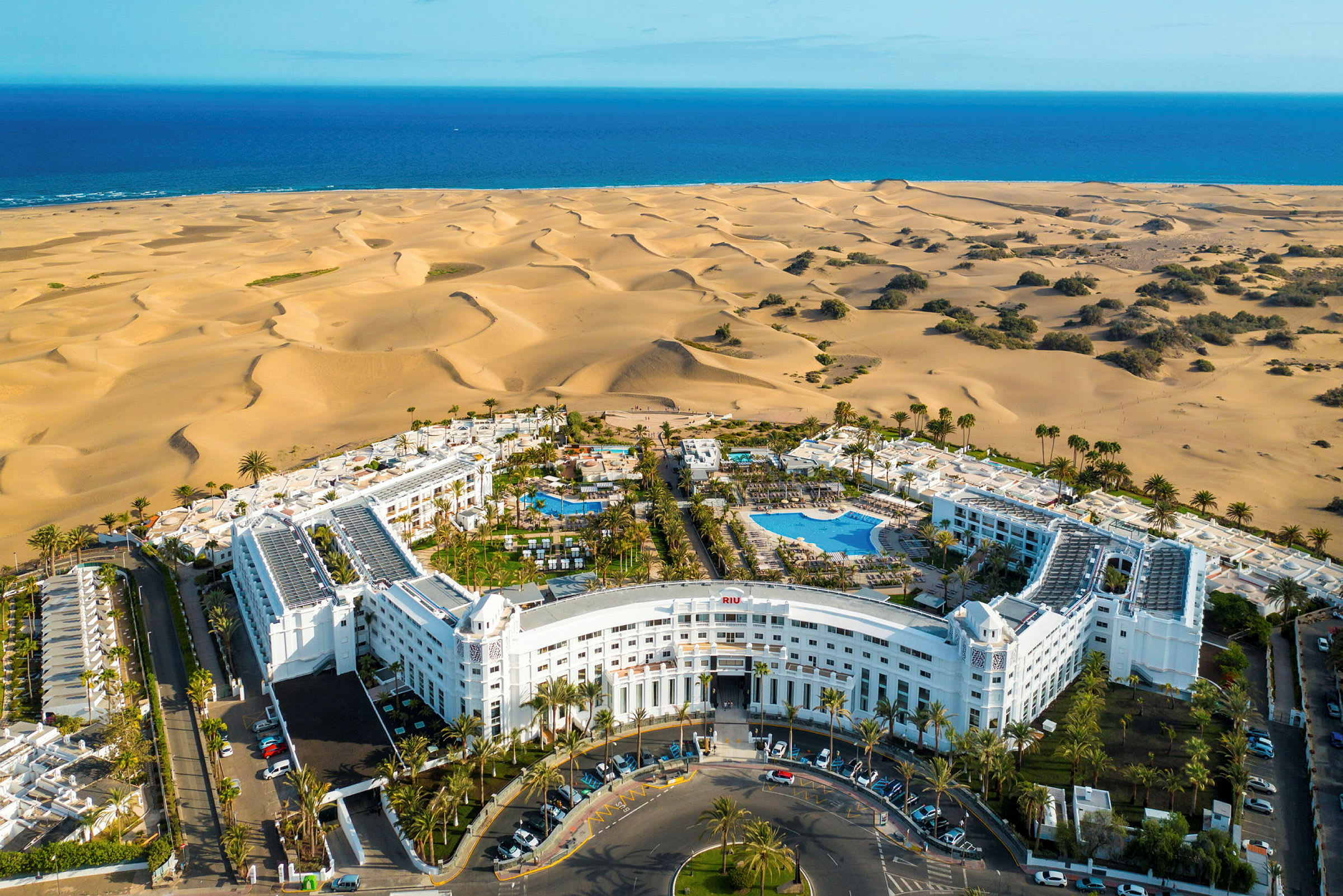 Luftfoto af et hotelkompleks med pools, omgivet af palmer og beliggende nær sandklitter og havet. (AI-genereret tekst)