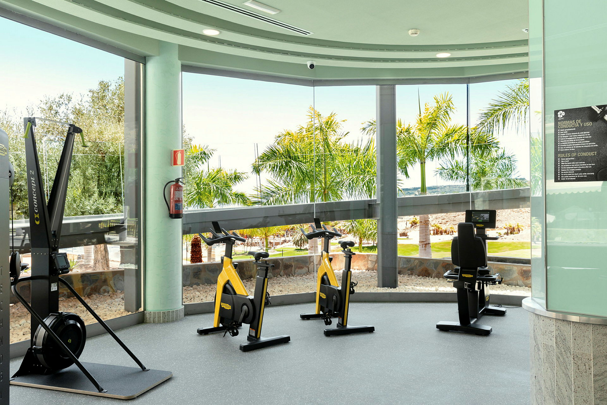 Et fitnesscenter med motionscykler og træningsudstyr, med store vinduer ud mod palmer og en have. (AI-genereret tekst)
