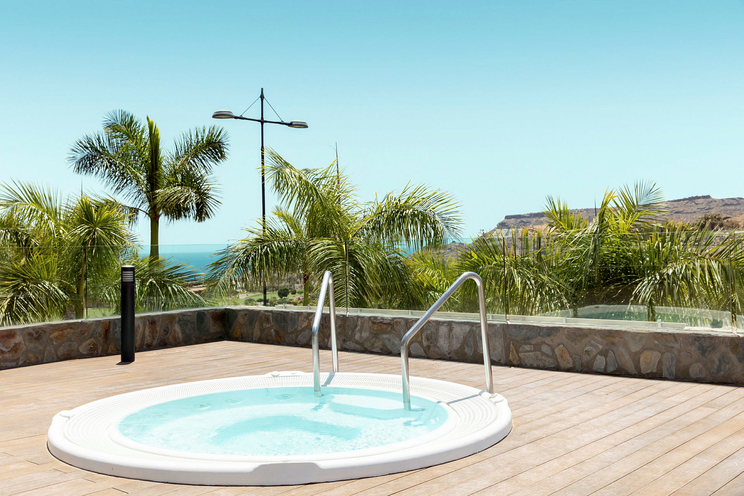 Rund jacuzzi på en terrasse med havudsigt og palmer. (AI-genereret tekst)