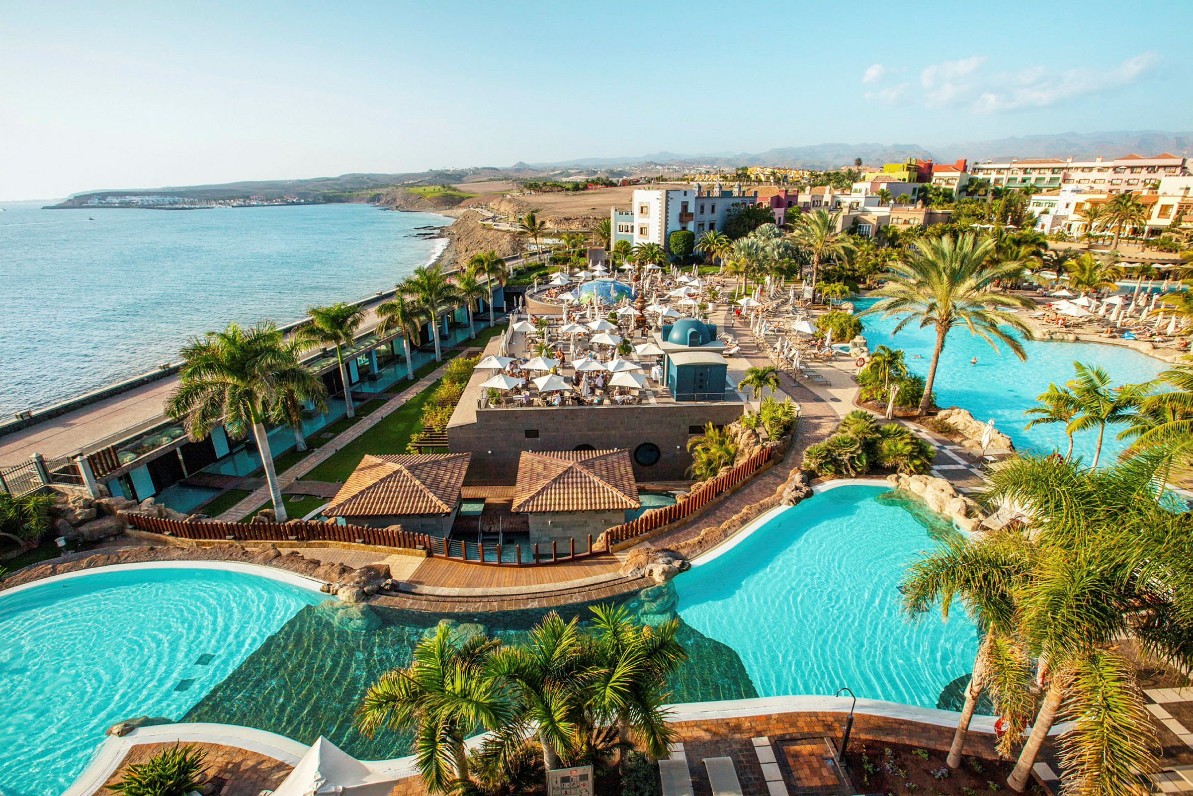 Udsigt over et feriehotel med swimmingpools, palmer og hav i baggrunden. (AI-genereret tekst)