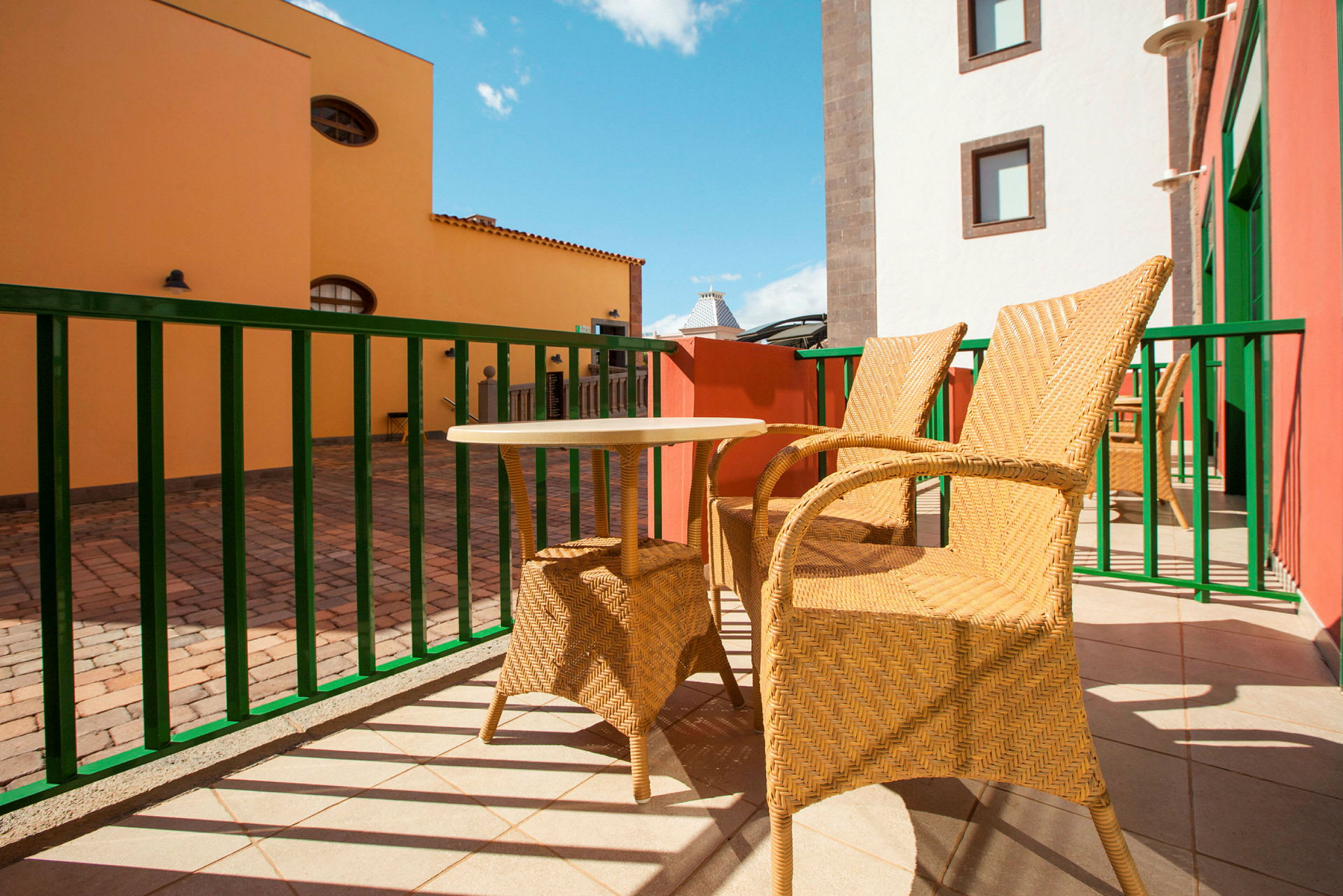 To flettede stole og et lille bord står på en solrig terrasse med farverige bygninger i baggrunden. (AI-genereret tekst)