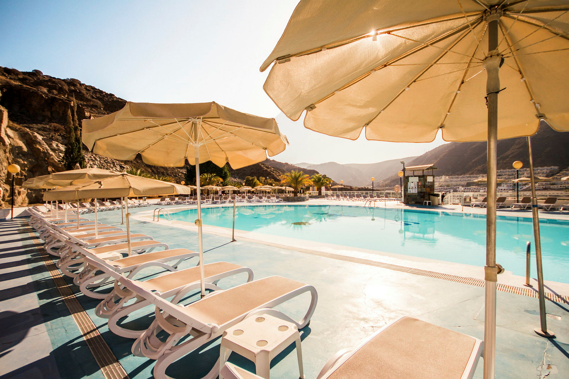 Udendørs swimmingpool med liggestole og parasoller, omgivet af bjerge. (AI-genereret tekst)