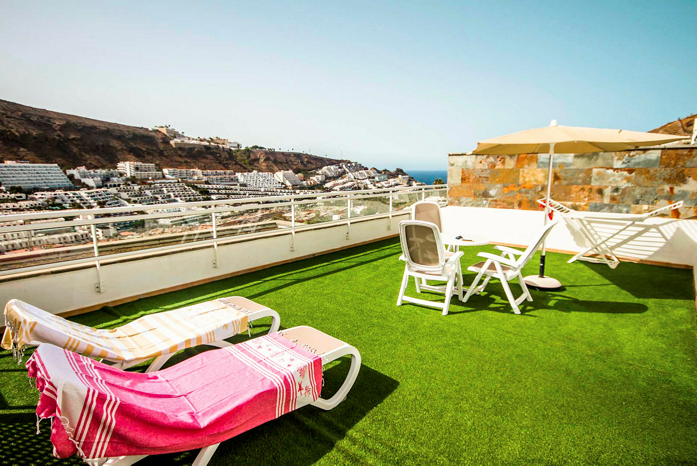 En terrasse med græsplæne, liggestole og parasol med udsigt over havet og omkringliggende bygninger. (AI-genereret tekst)
