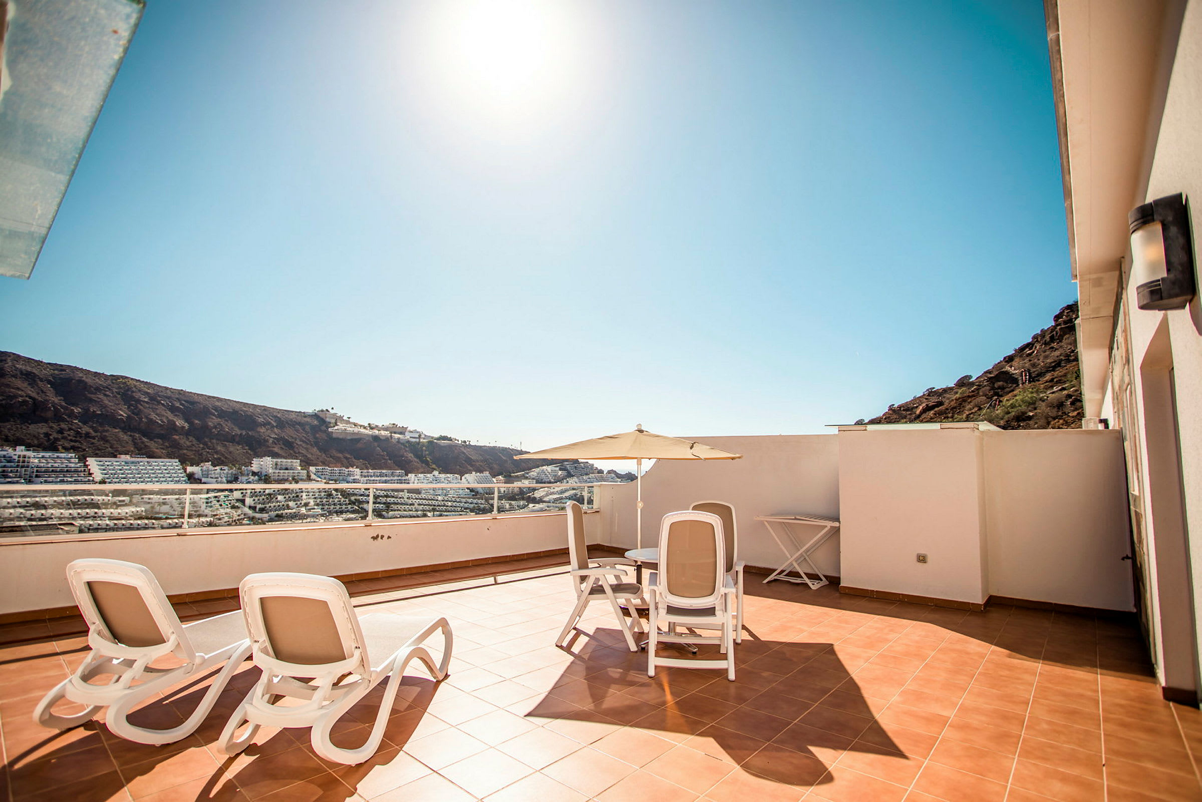 Solrig terrasse med havemøbler og udsigt til omgivelser med bygninger og bjerge. (AI-genereret tekst)