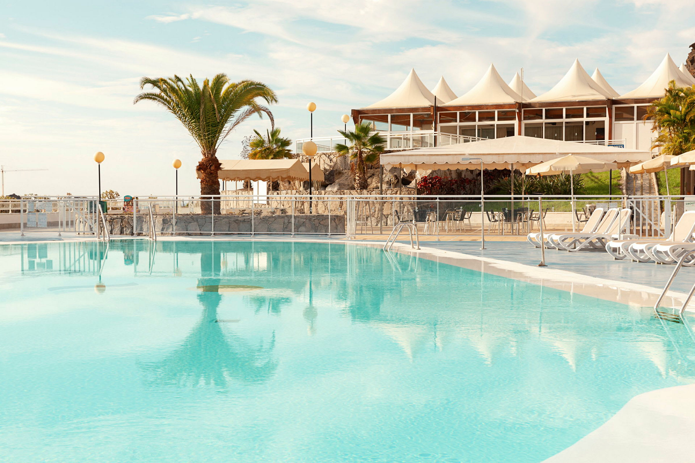 En udendørs swimmingpool med liggestole og udsigt til en bygning med hvide teltformede tage. (AI-genereret tekst)