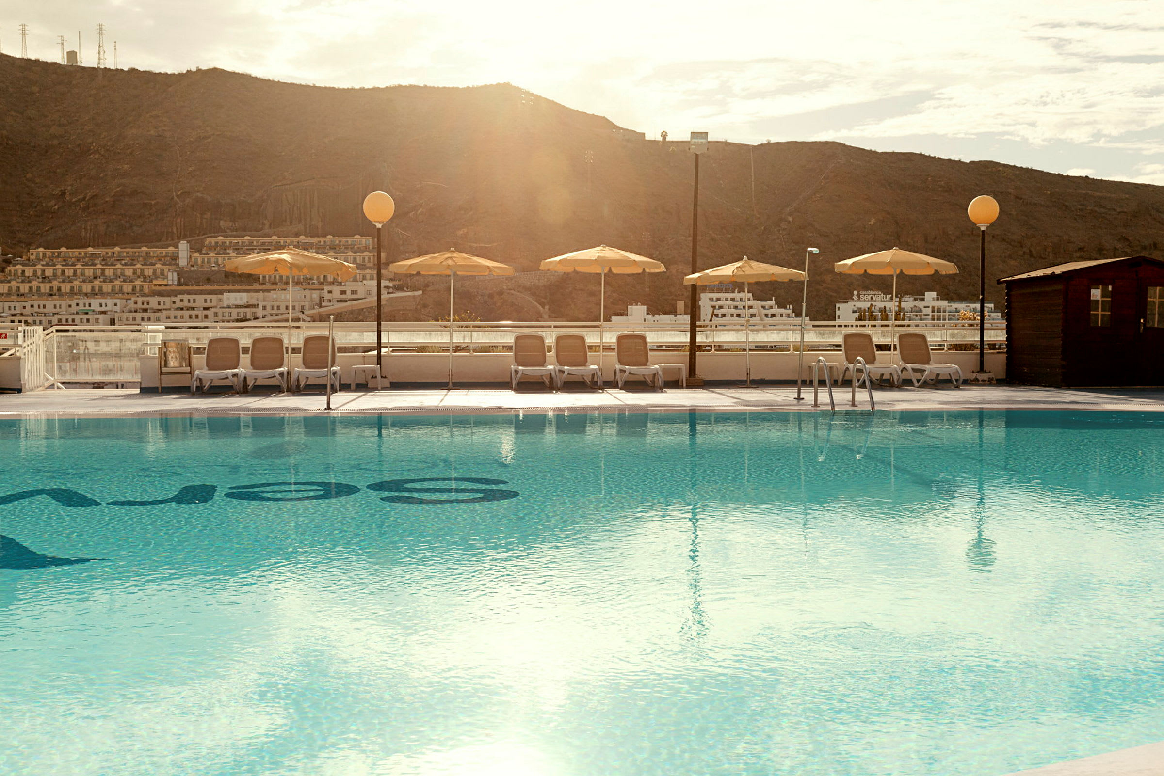 En udendørs swimmingpool med liggestole og parasoller i baggrunden. (AI-genereret tekst)