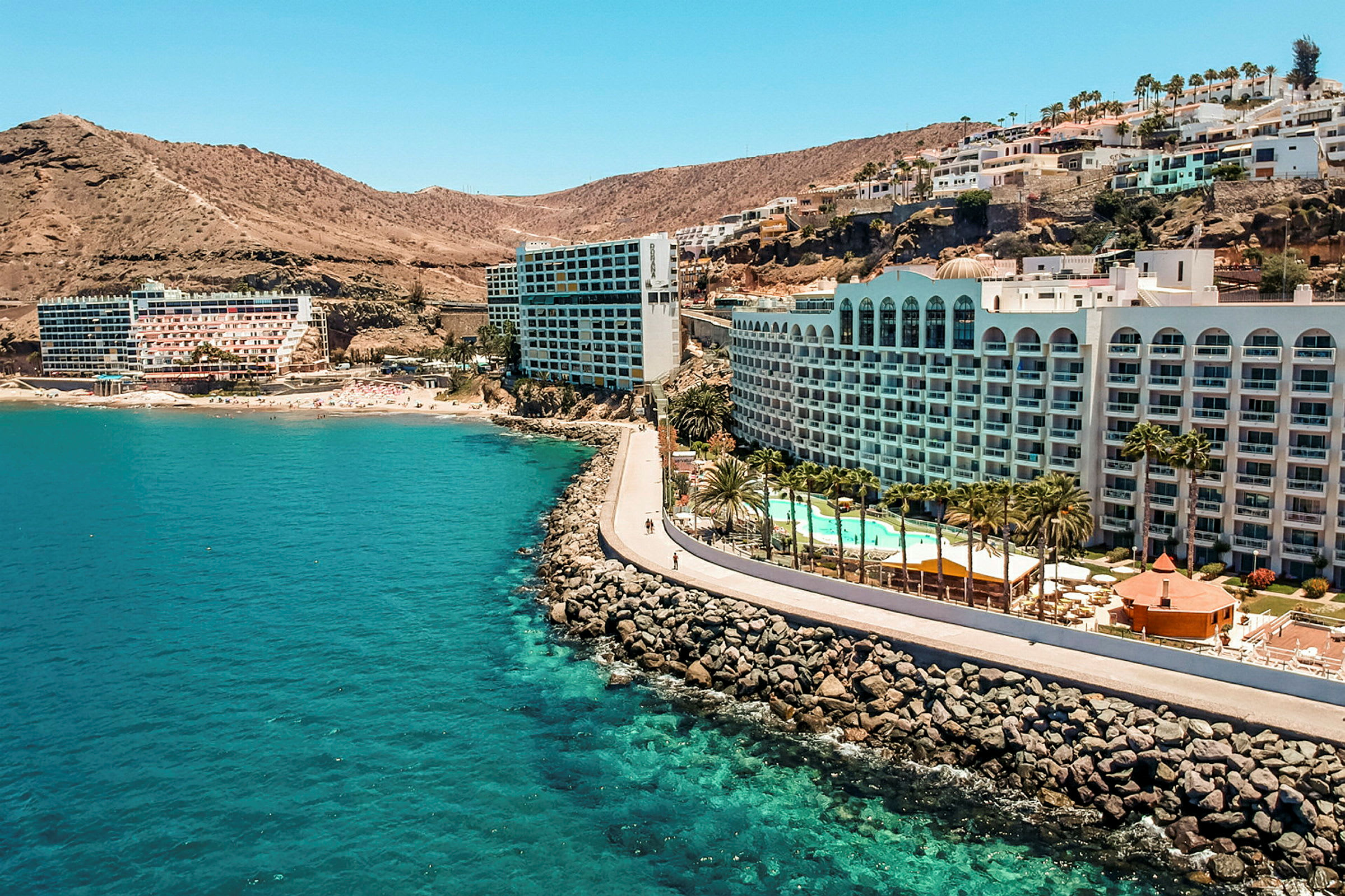 Hotel ved en kyst med klipper, pool og palmer. (AI-genereret tekst)