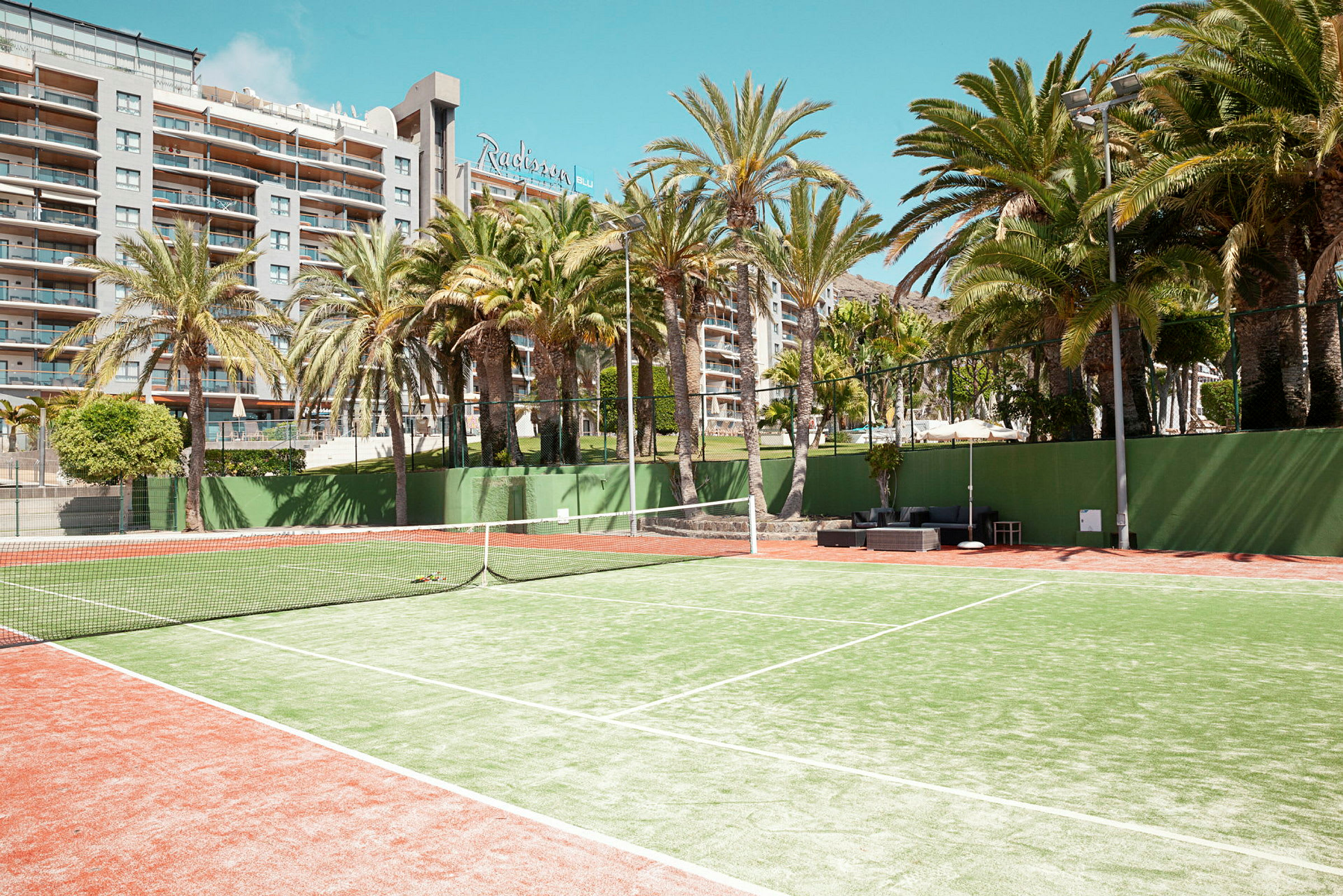 Tennisbane med grusunderlag omgivet af palmer, med et hotel i baggrunden. (AI-genereret tekst)