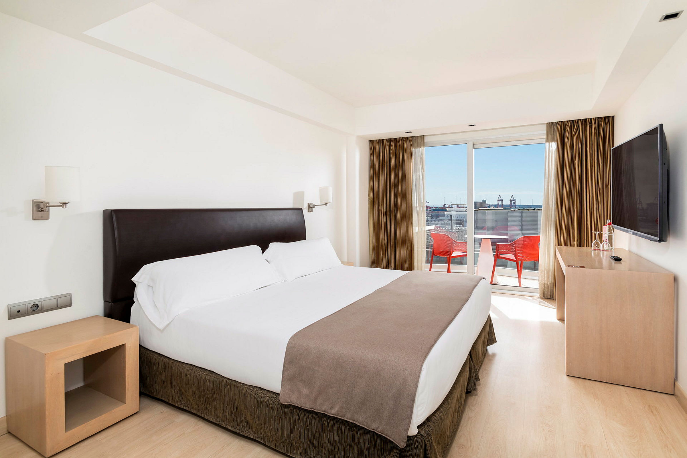 Hotelværelse med en dobbeltseng, sengebord og TV. Vindue med udsigt til en balkon med stole og bord. (AI-genereret tekst)
