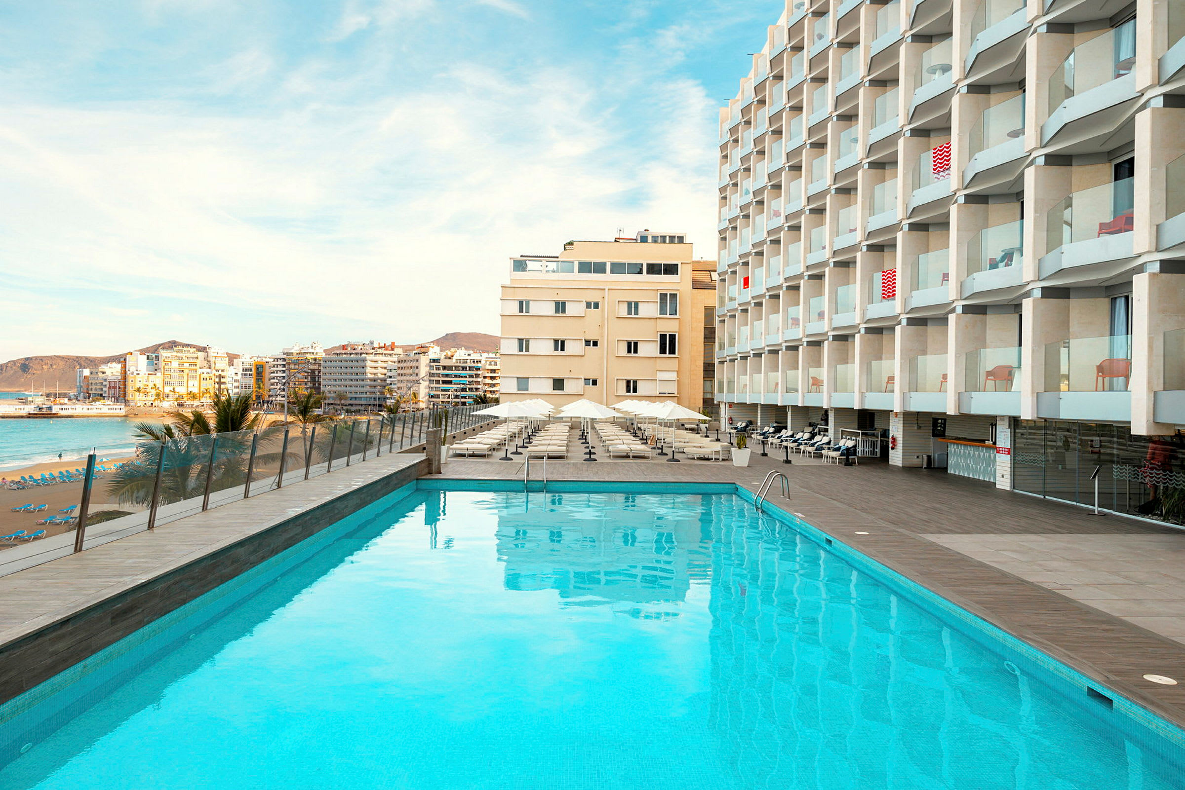 Udendørs pool foran hotelbygning, med strand og by i baggrunden. (AI-genereret tekst)