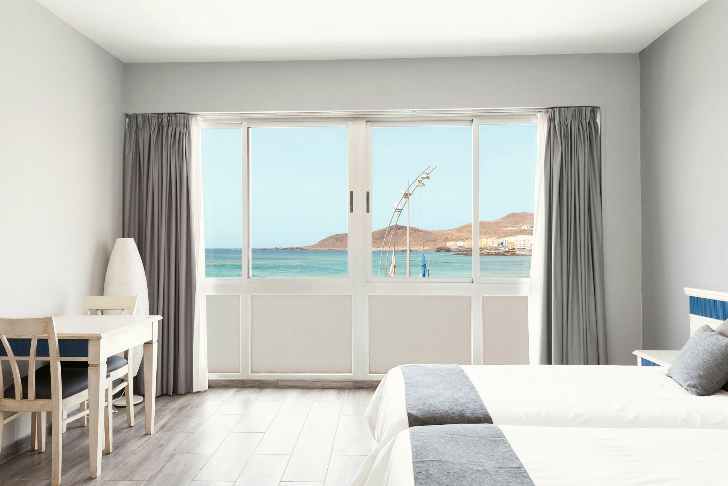Lyst hotelværelse med udsigt over havet gennem en stor vinduesfront. (AI-genereret tekst)